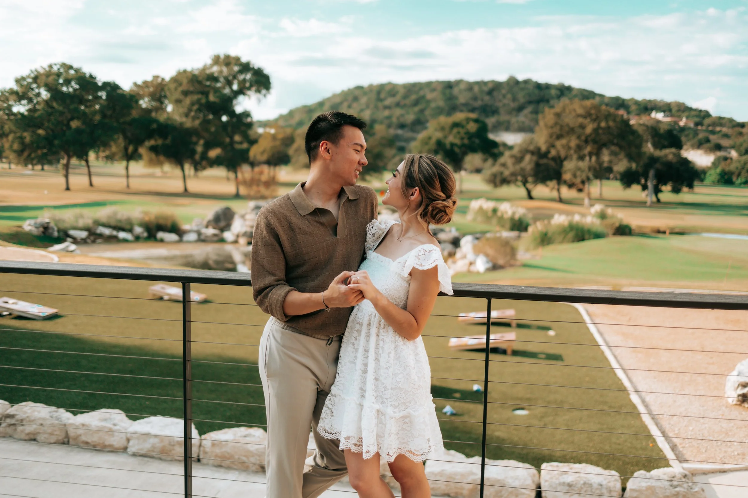 A couple stands on a balcony overlooking a golf course with trees and hills in the background, holding hands and gazing at each other.