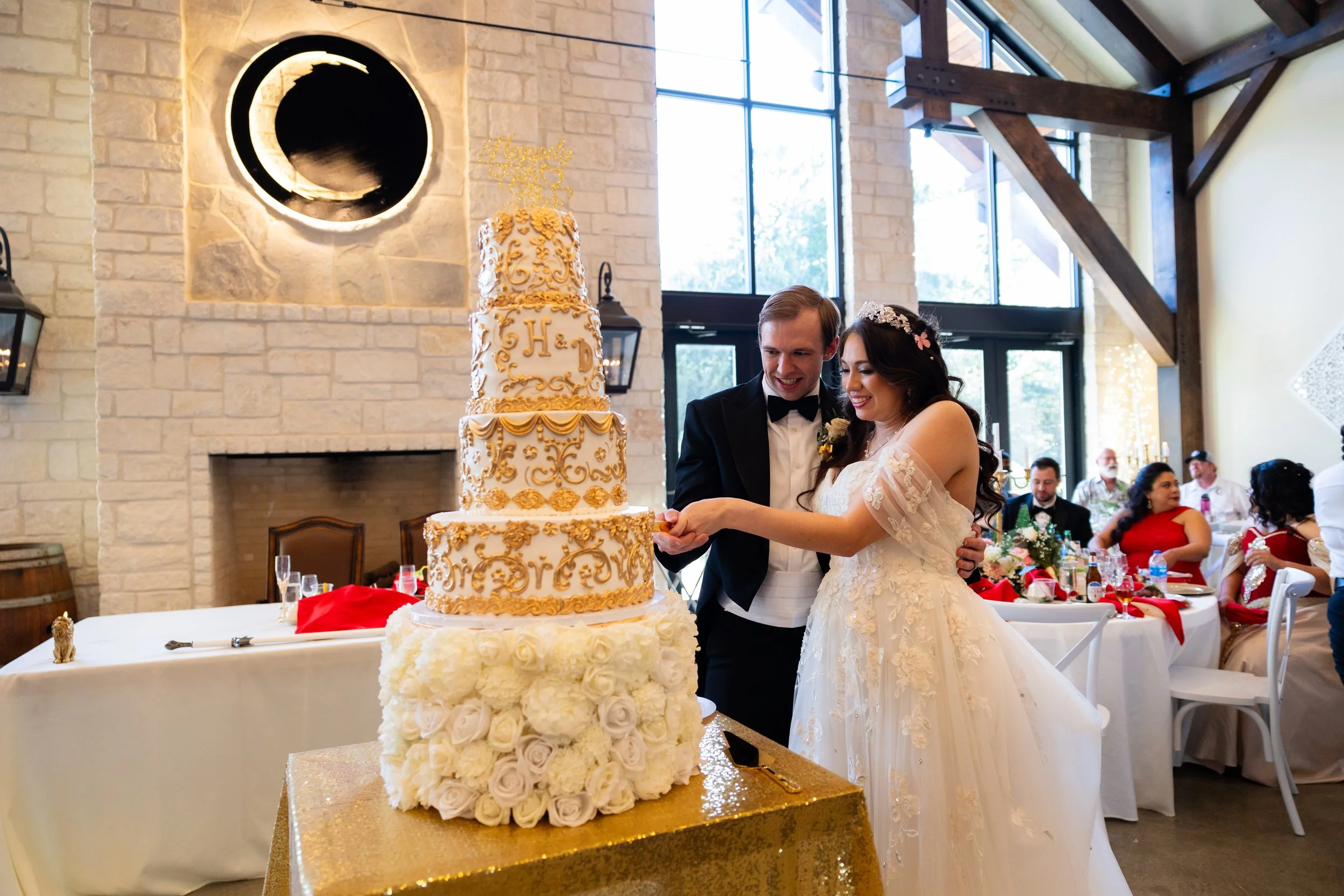 A bride and groom cutting their wedding cake at a reception, surrounded by guests seated at decorated tables.