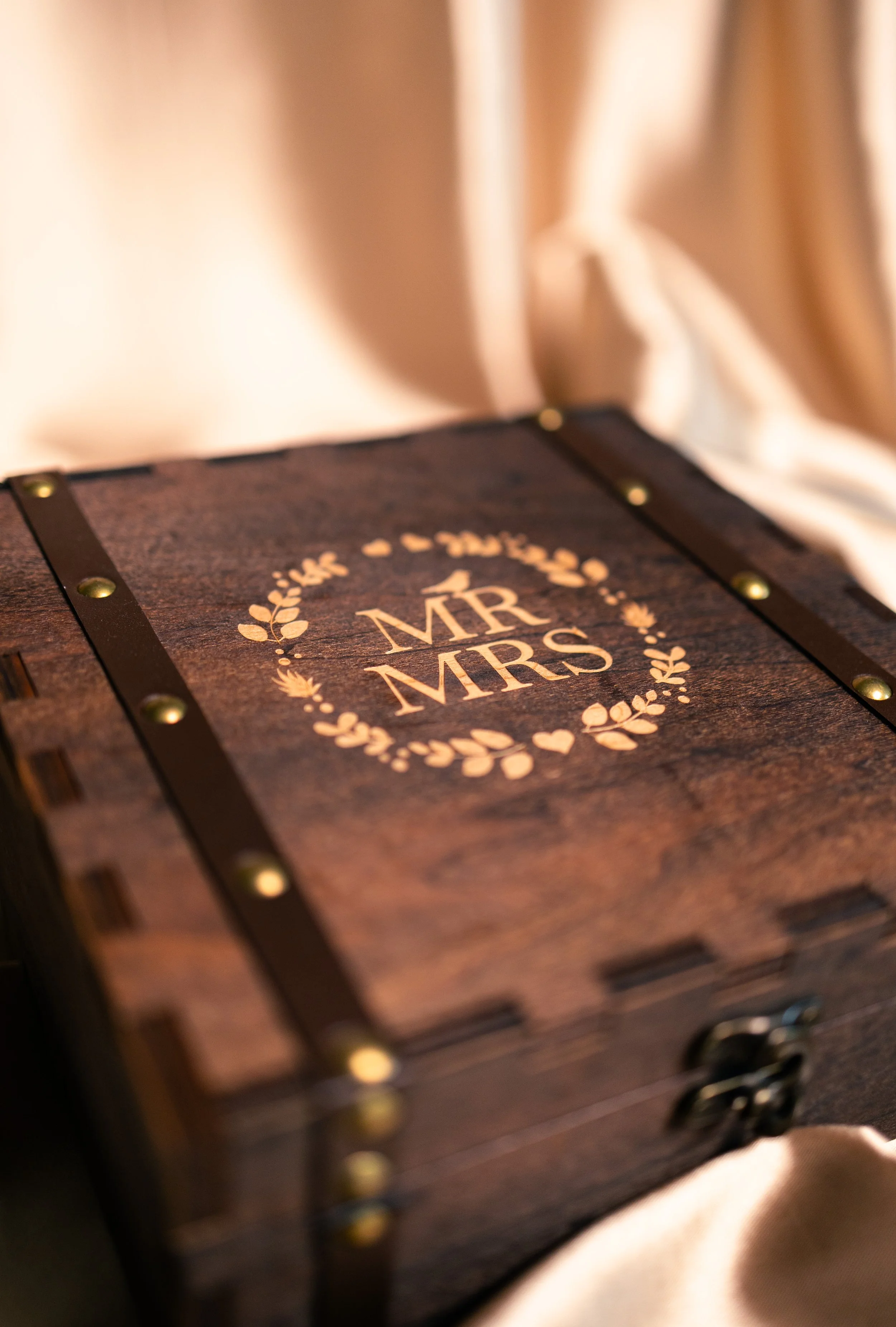 A wooden box with the words 'MR MRS' in the center, surrounded by a decorative wreath of leaves and hearts, resting on a beige fabric background.