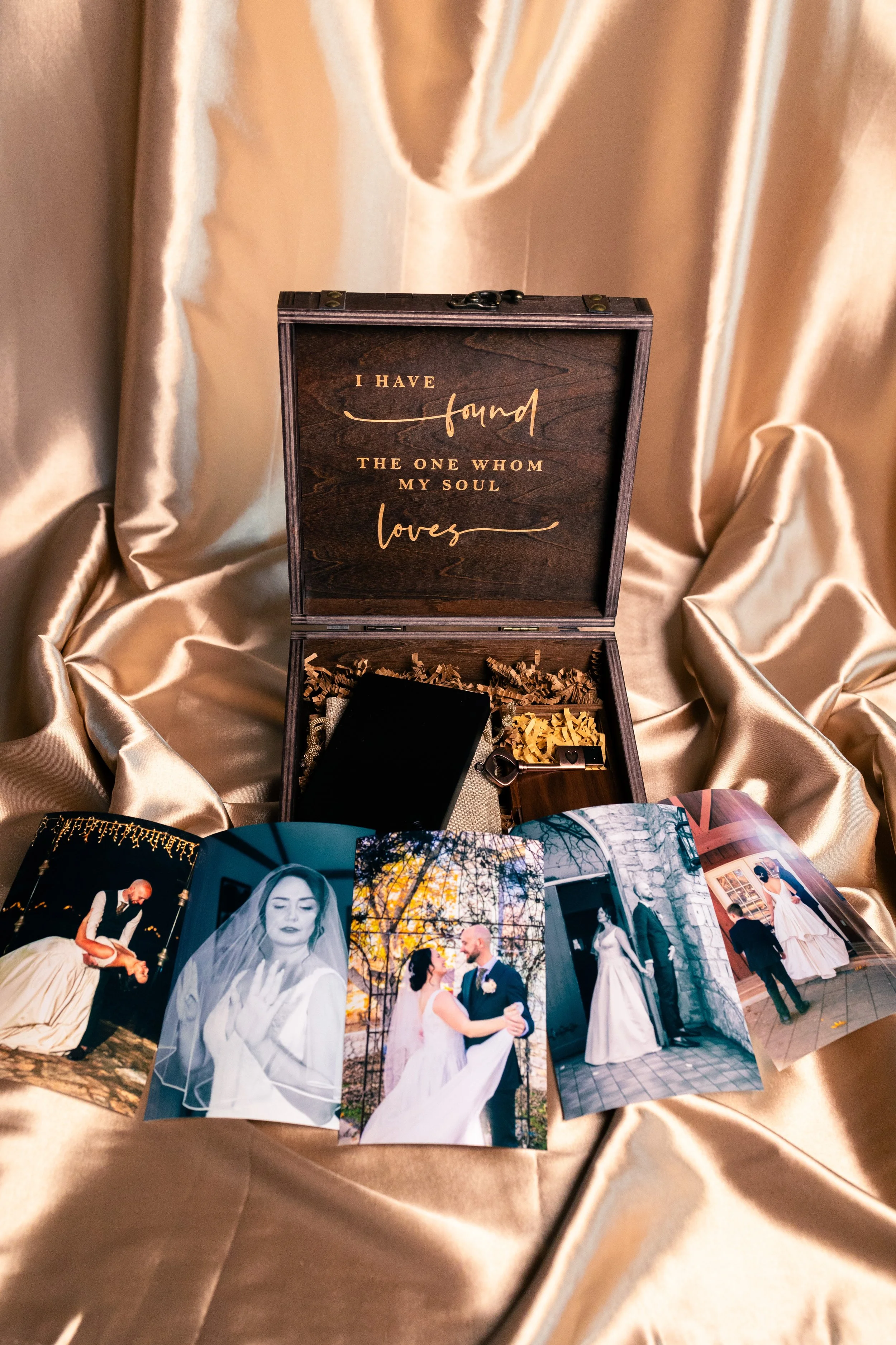 A photo of a wedding keepsake box with a golden quote inside the lid that says, 'I have found the one whom my soul loves,' surrounded by wedding photos of a couple on a satin fabric background.