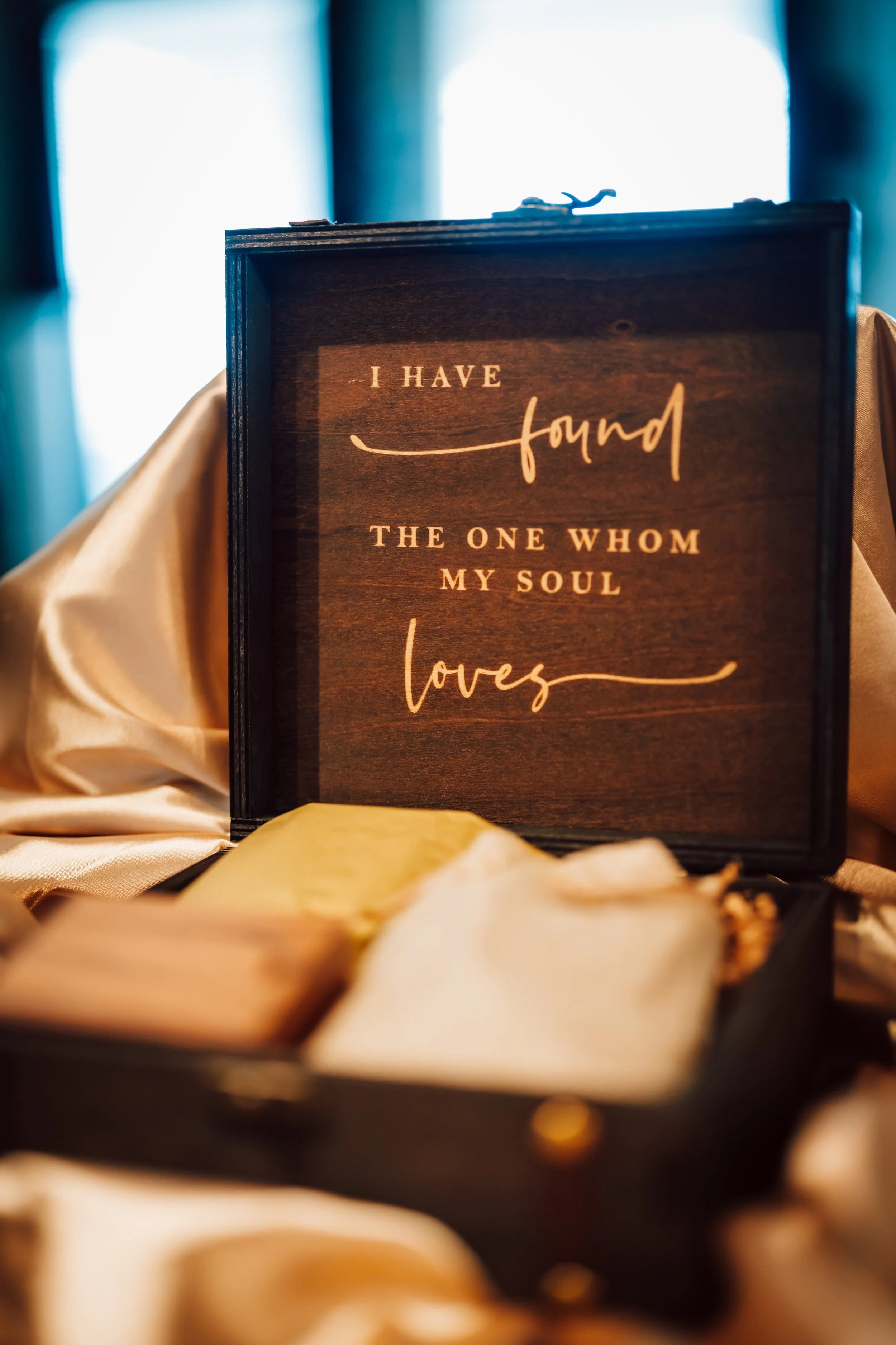 A wooden box with a quote inside that reads, 'I have found the one whom my soul loves.' The box is surrounded by silk fabric and what appears to be jewelry, with a bright light in the background.