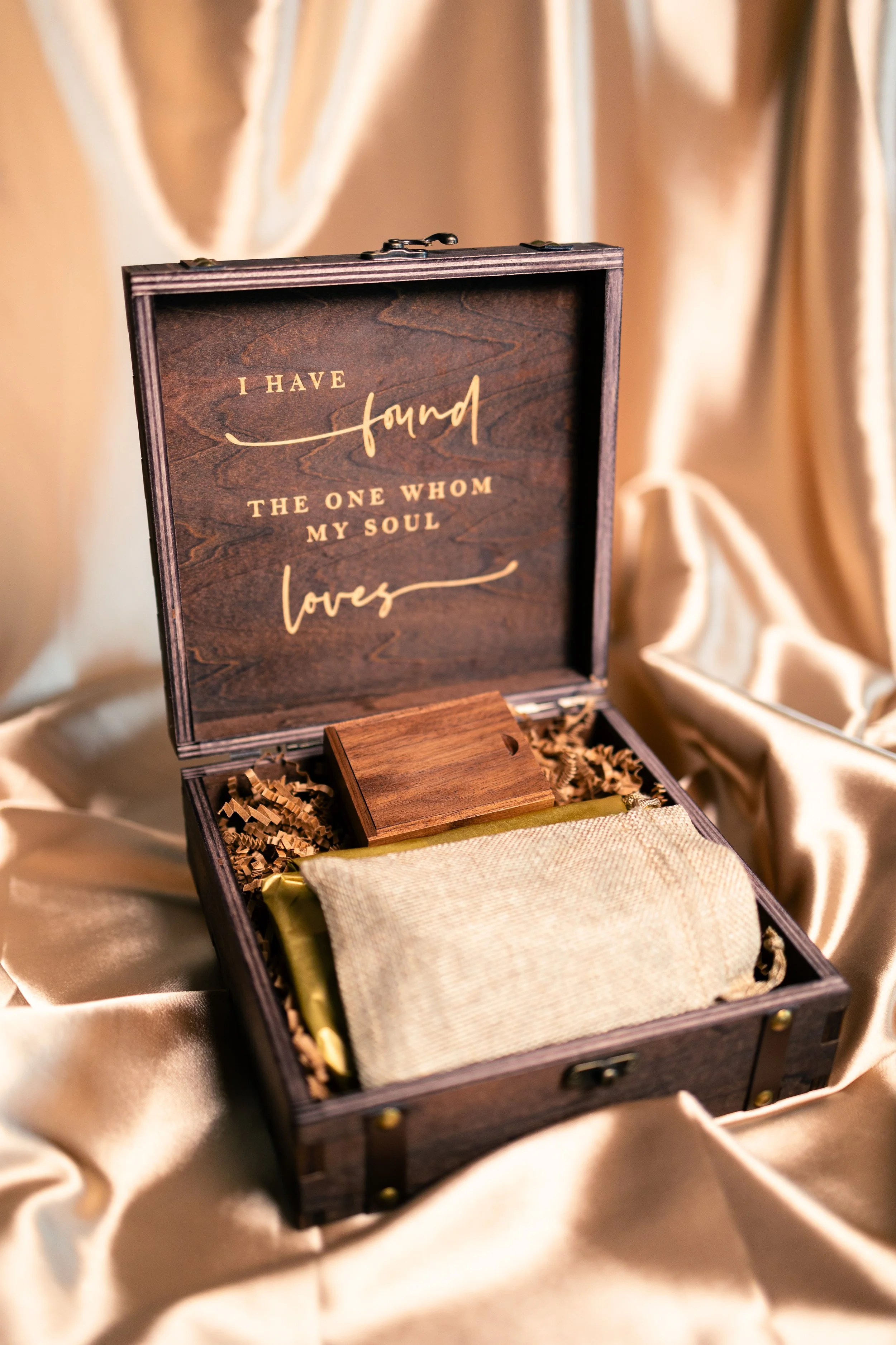 A wooden box with a hinged lid, with gold lettering inside the lid that reads, "I have found the one whom my soul loves." The box contains a wooden stamp and some wrapped items, set on satin fabric.