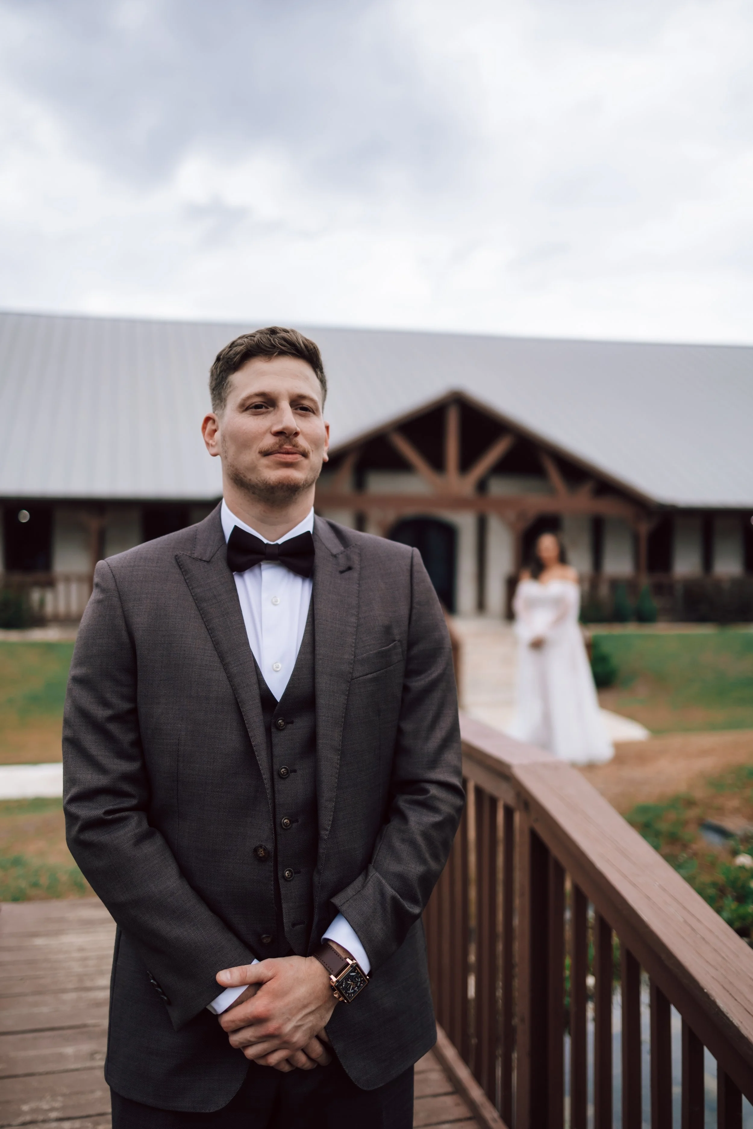 A man in a dark grey suit, white shirt, and black bow tie stands on a wooden deck outside a building with a grey metal roof. In the background, a woman in a white wedding dress is slightly out of focus.