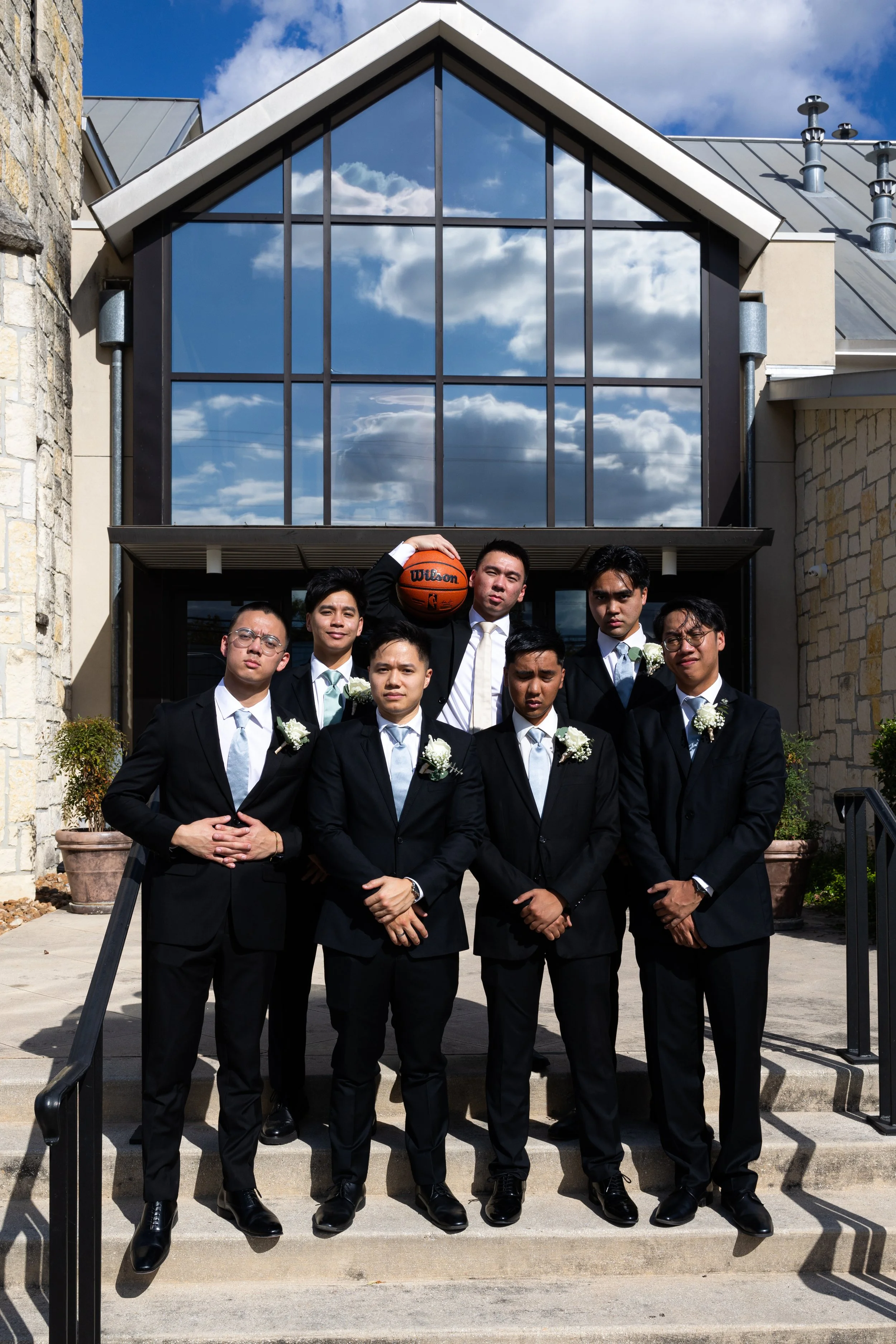 Group of seven men in formal suits standing on outdoor stairs in front of a building with reflective glass windows, with one holding a basketball overhead.