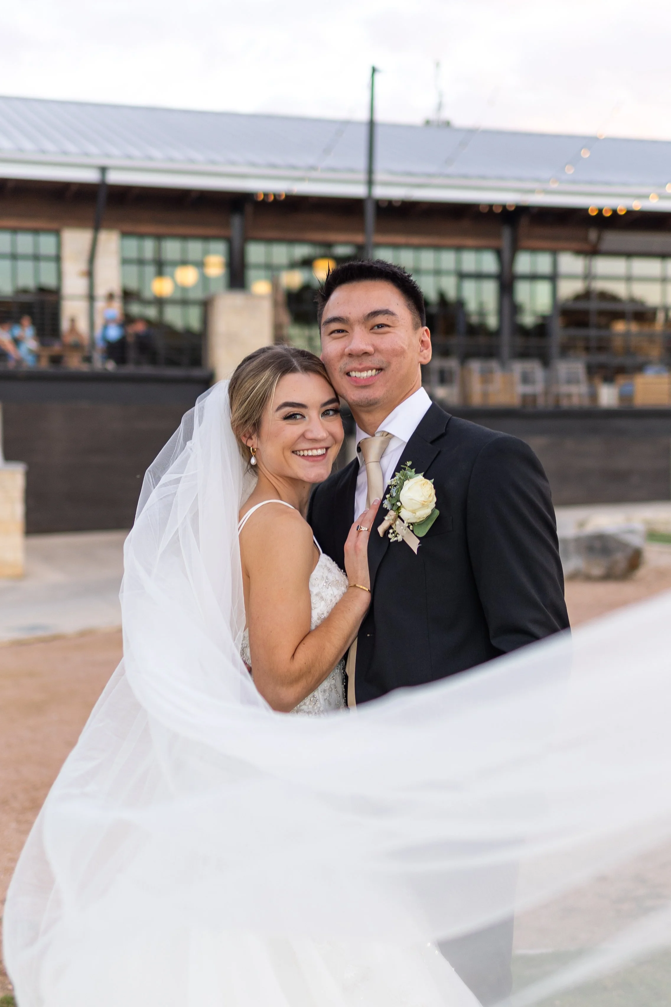 A bride and groom smiling closely together, outdoors during a wedding, with the bride's veil flowing around them.
