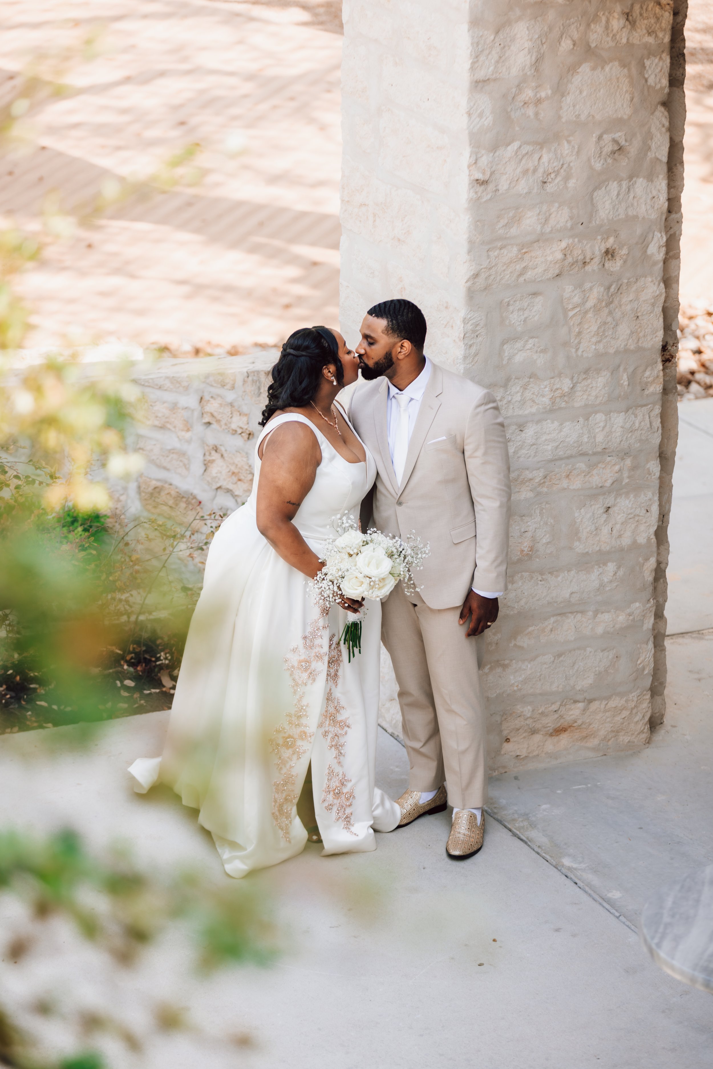 A couple dressed in wedding attire sharing a kiss outside next to a stone pillar, with the bride holding a bouquet of white flowers.