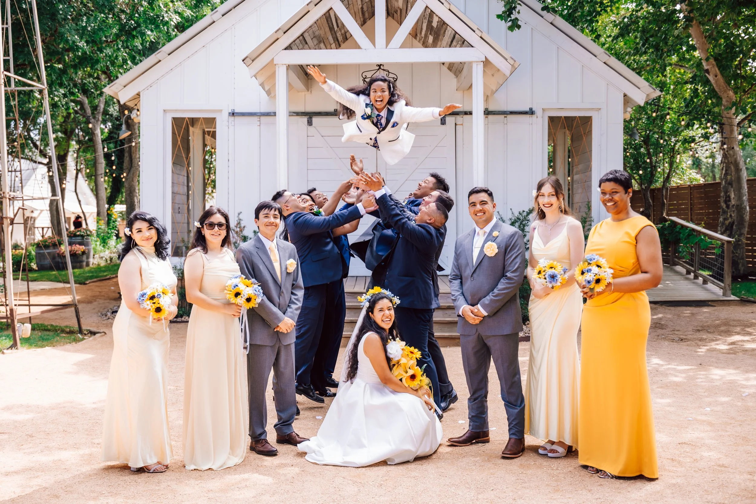 A wedding party celebrating outdoors in front of a small white chapel, with the bride, groom, bridesmaids, and groomsmen. The groom and groomsmen are throwing the bride into the air, and she is smiling happily. The group is dressed in formal attire, 