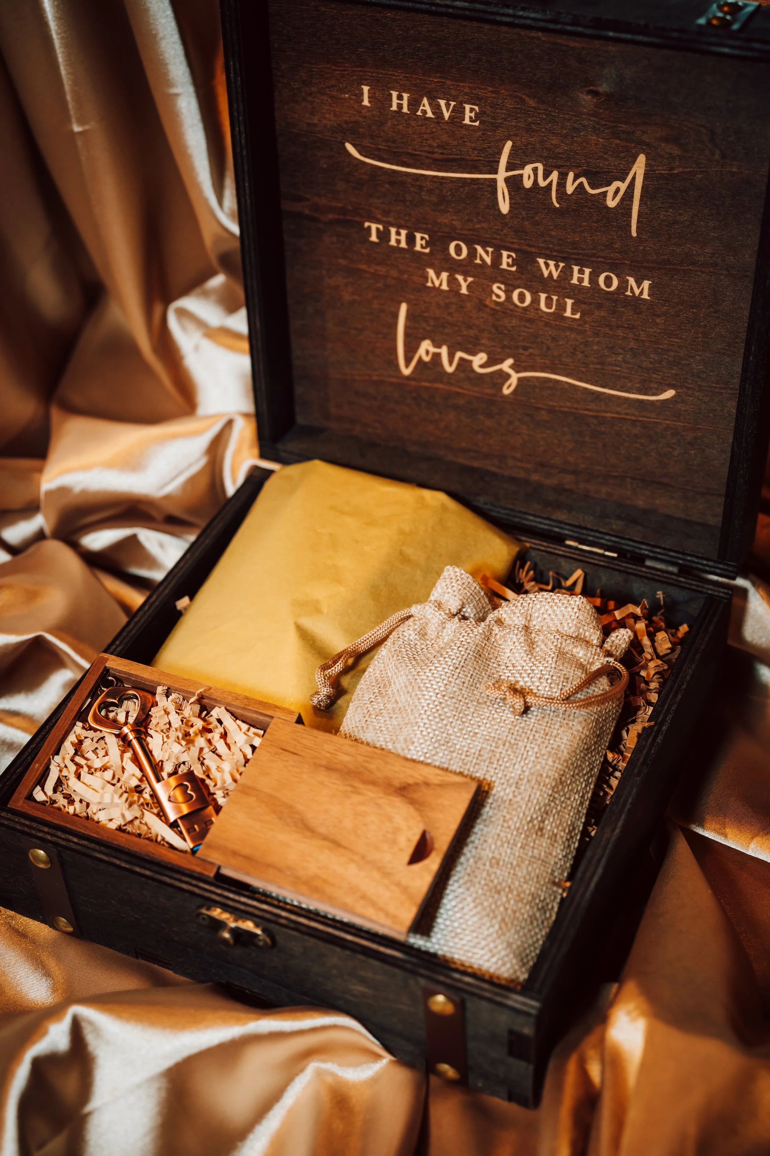 A gift box with a heartfelt quote on the inside lid, containing a small burlap bag, a wrapped item, a wooden box, and a key, surrounded by gold satin fabric.