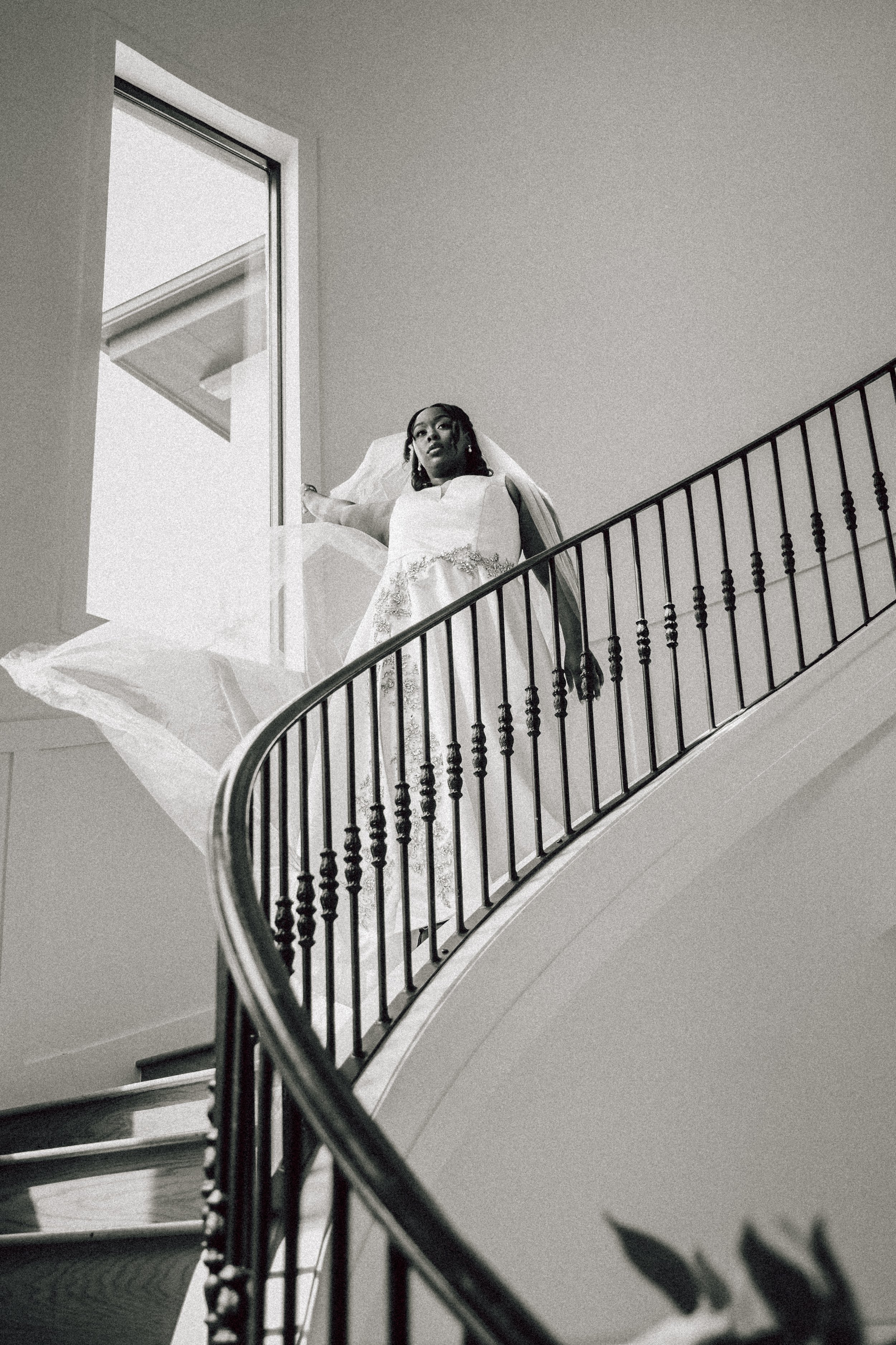 A woman in a wedding dress standing on a staircase landing near a window, looking down towards the camera with a contemplative expression.