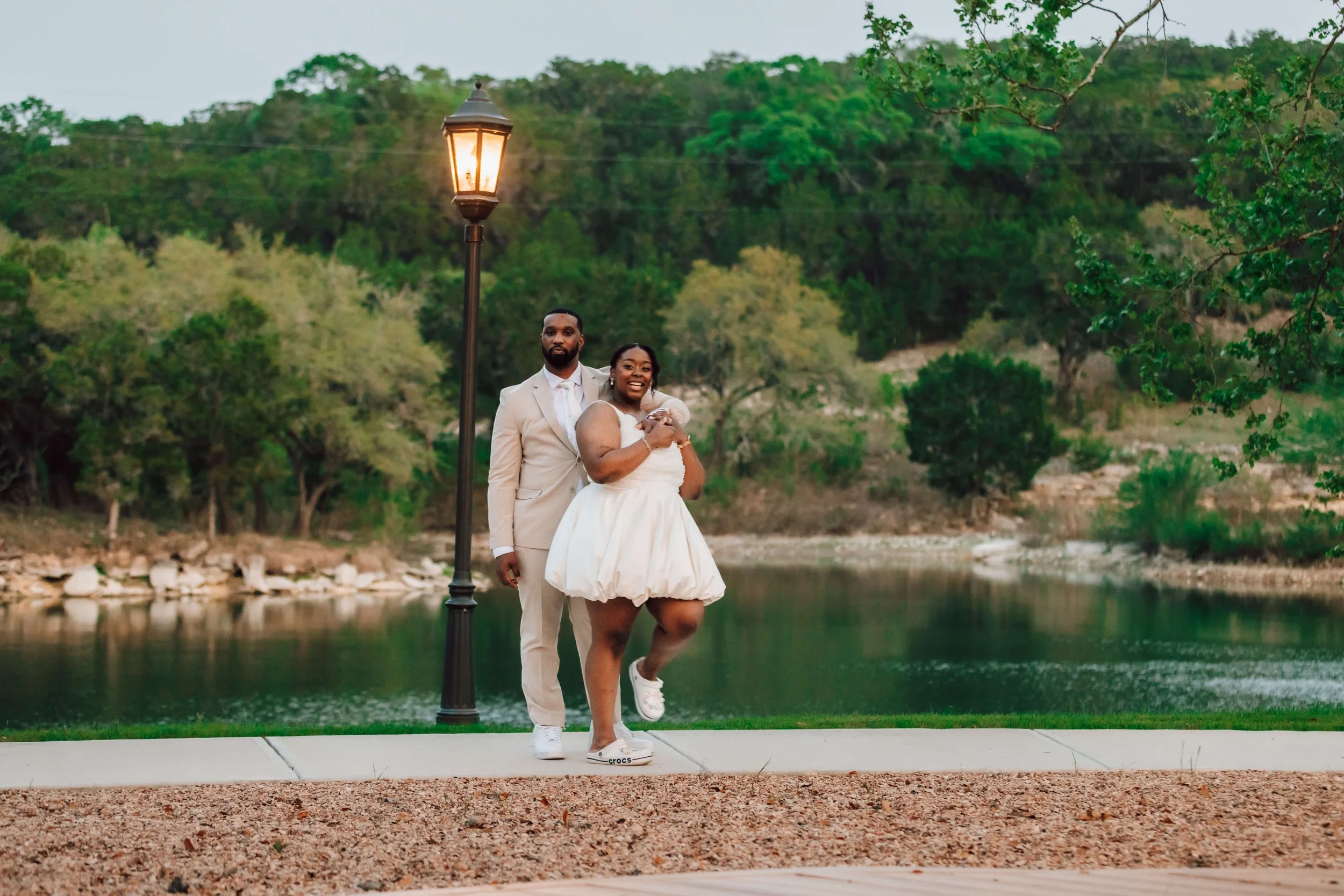A happy couple dressed in wedding attire standing beside a lamppost by a river, with greenery and trees in the background during daytime.