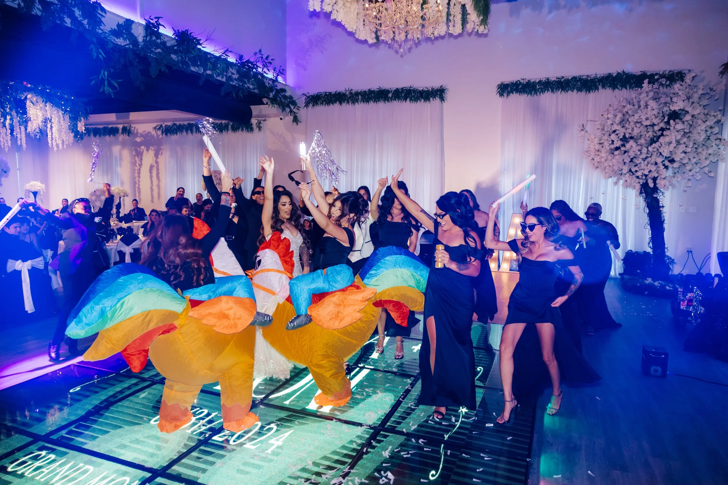 People dancing at a celebration, with some dressed in colorful costumes resembling llamas or alpacas, surrounded by festive decorations and floral arrangements.