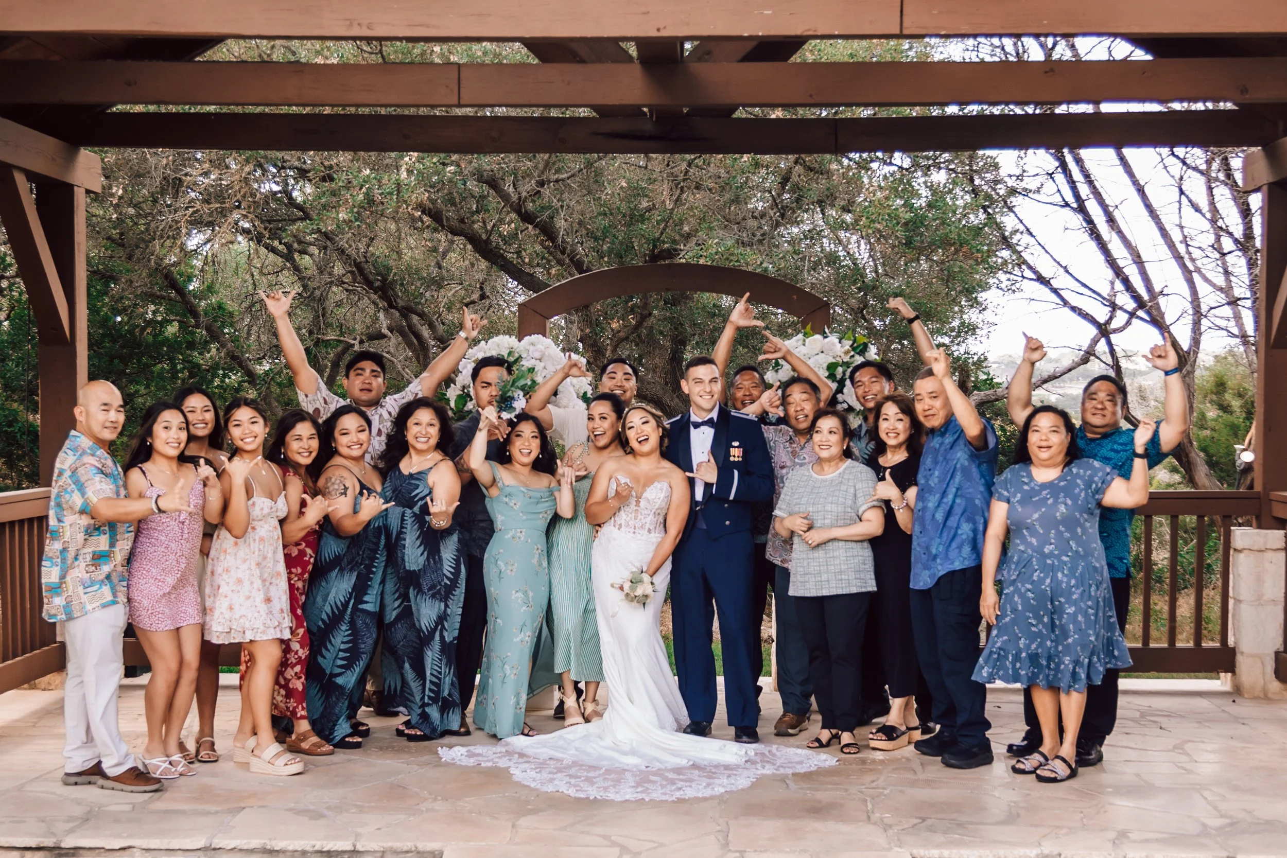 Group of people at a wedding party outdoors under a wooden structure, with trees in the background. The bride and groom are at the center, smiling and celebrating with family and friends.