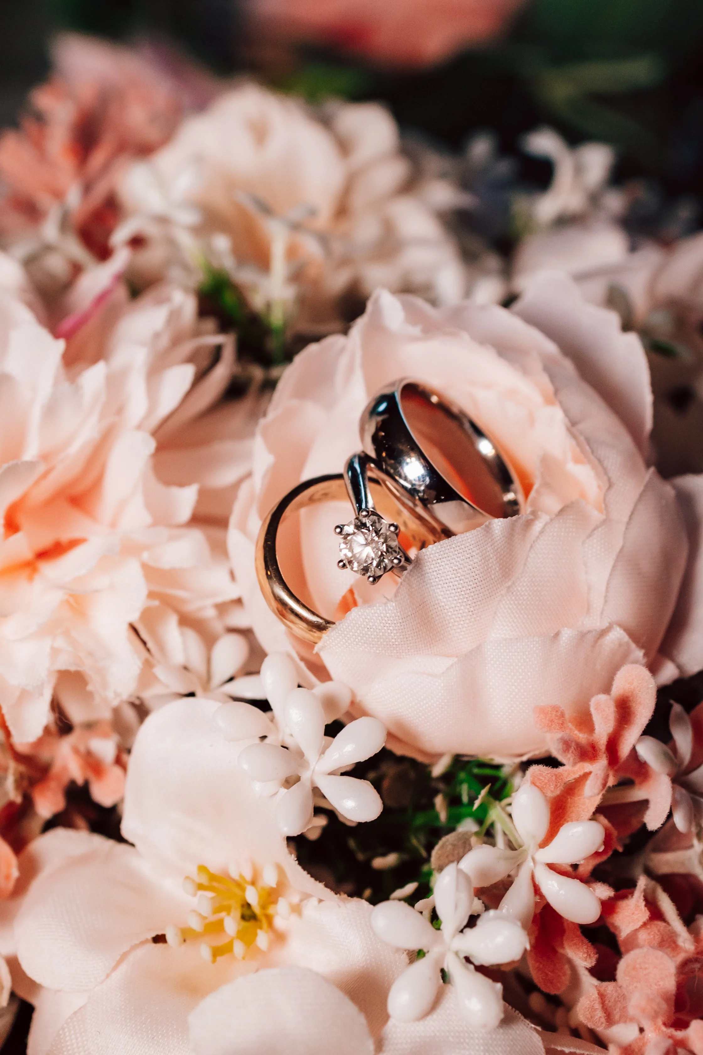 Close-up of wedding rings, one with a diamond solitaire, placed on pink and white artificial flowers.