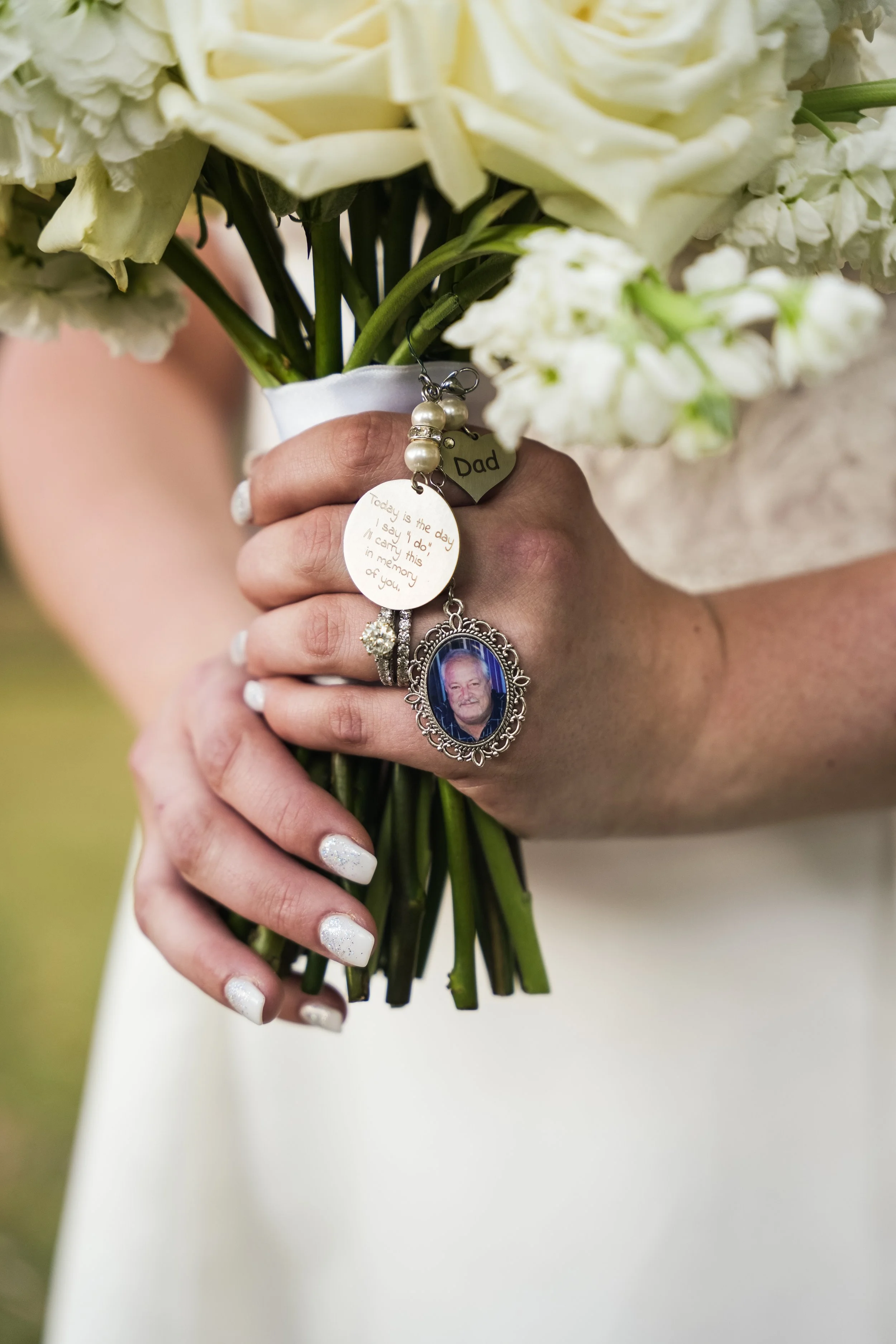 Close-up of a woman's hand holding a bouquet of white roses, with a wedding ring, decorated with charms including a photograph of a man, a pearl, and a tag labeled 'Dad'.