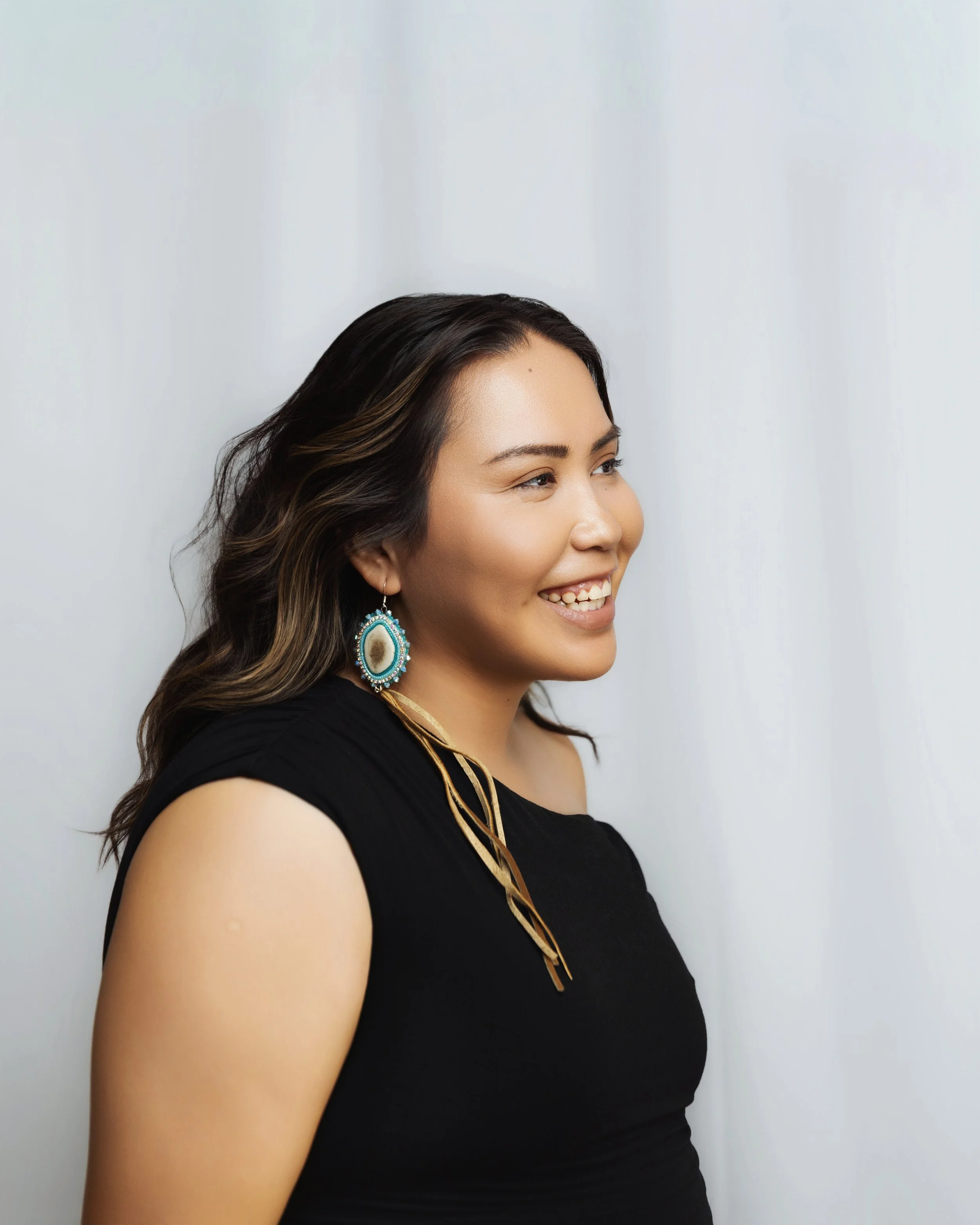 Smiling woman with dark hair, wearing earrings and a black sleeveless top, standing against a plain light-colored background.