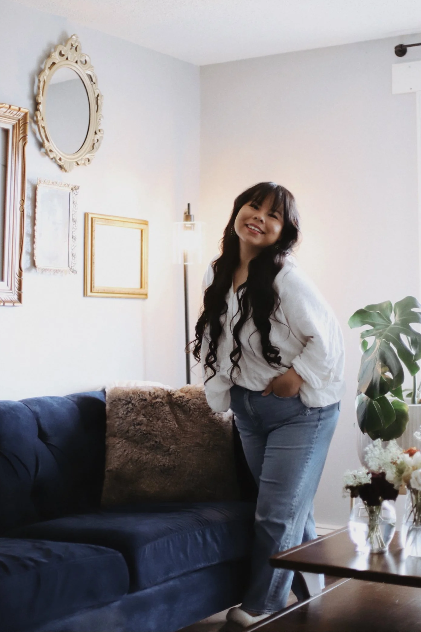 A woman with long dark hair, wearing a white shirt and light blue jeans, standing next to a navy blue sofa with a fluffy brown pillow, in a living room with framed artwork on the wall, a tall lamp, and a window with a curtain, smiling at the camera.