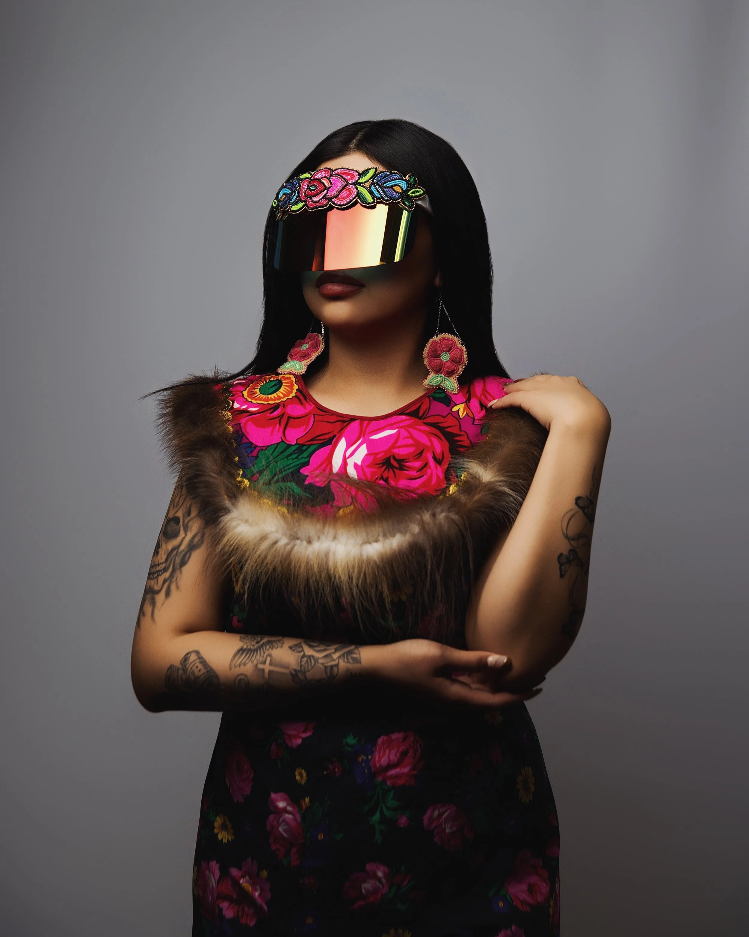 A woman with tattoos on her arms wearing a floral dress with a fur collar, large embroidered earrings, and a decorative visor sunglasses with floral embroidery, standing against a plain gray background.