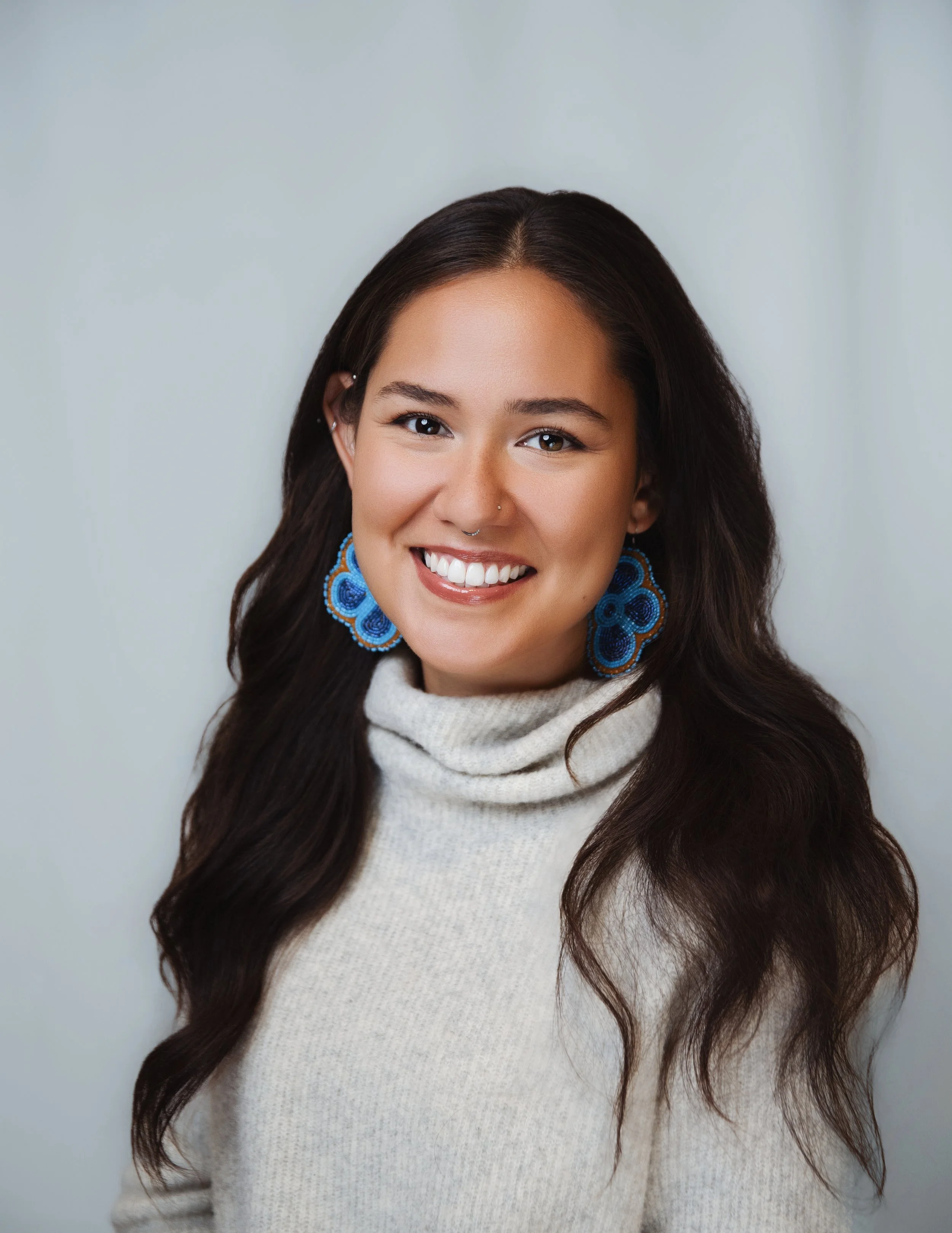 Portrait of a young woman with long dark hair, smiling, wearing blue earrings and a cream-colored turtleneck sweater, standing against a light background.