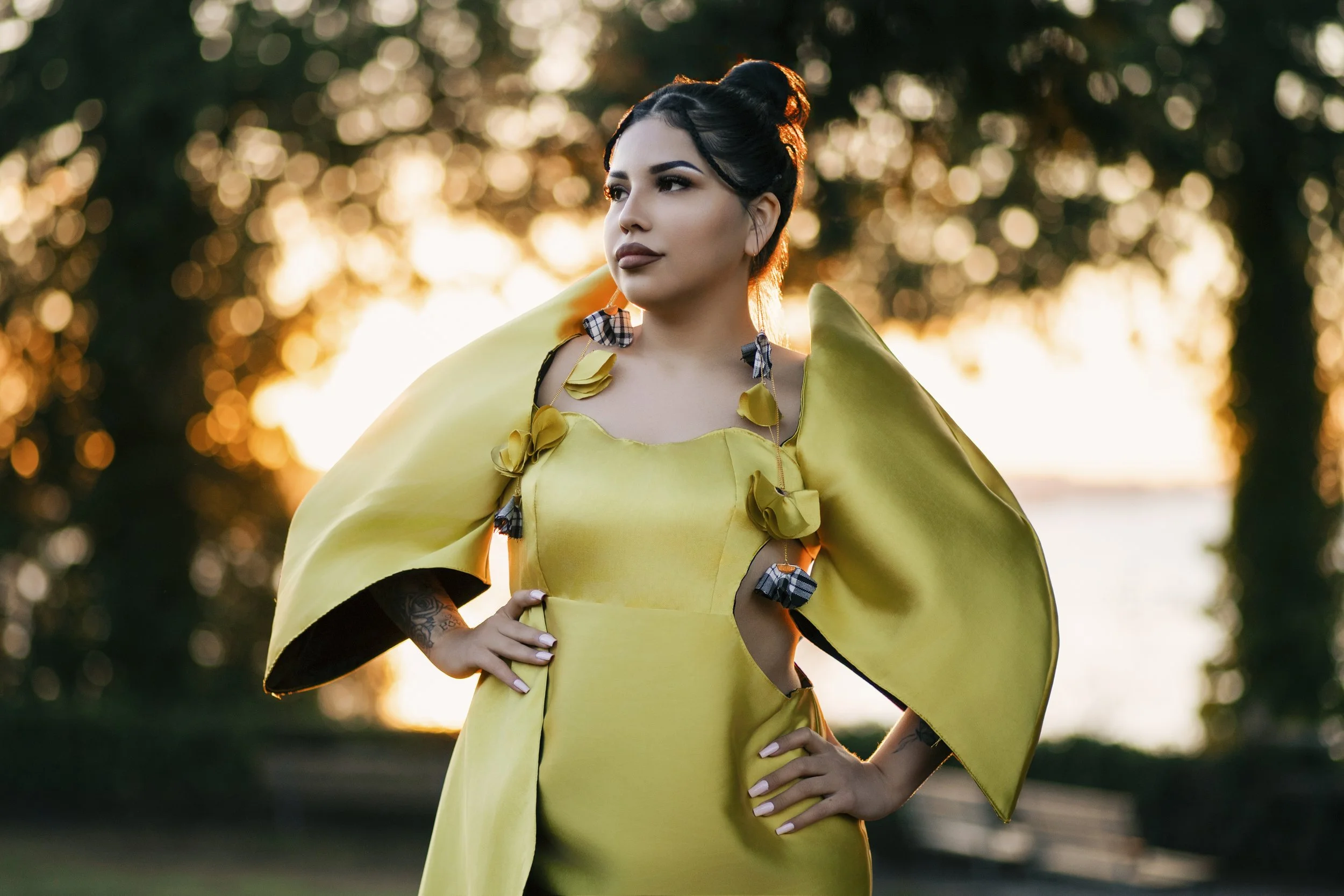 A woman in a yellow dress with unique cutouts and decorative fabric flowers, standing outdoors at sunset with trees in the background.