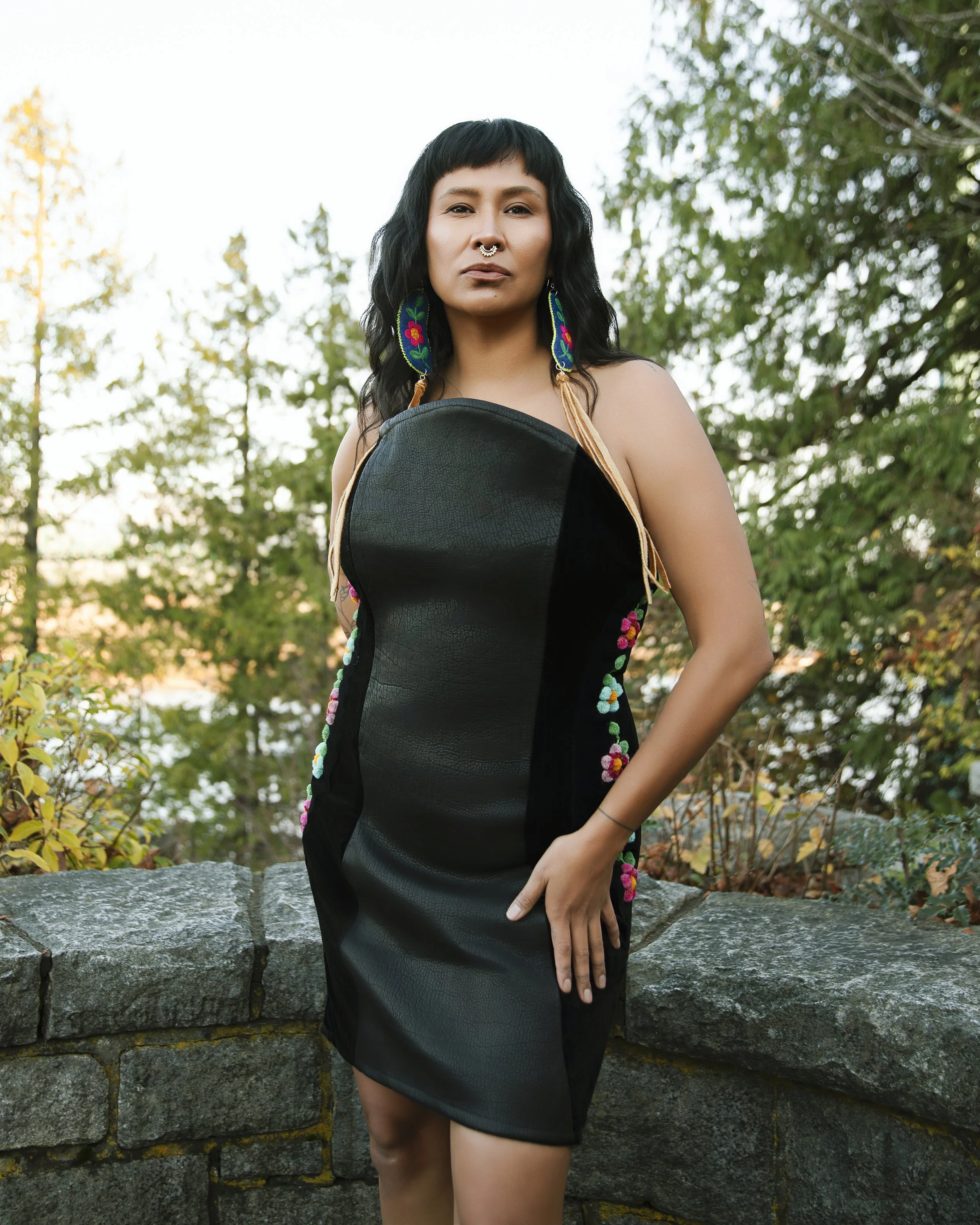 Woman wearing a black dress with colorful embroidery, standing outdoors near a stone wall, with trees in the background.