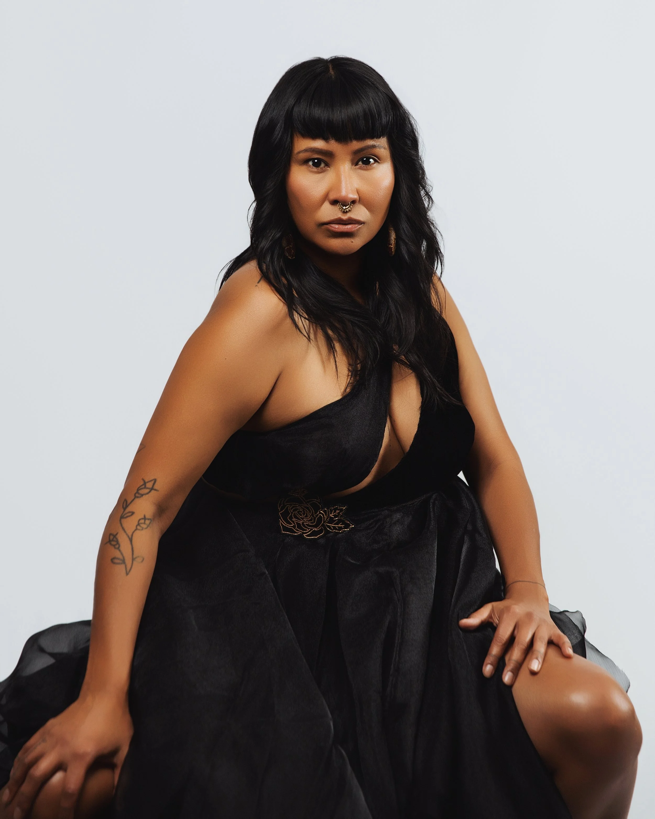 A woman with dark hair and a septum piercing wearing a black dress, sitting on a white background.