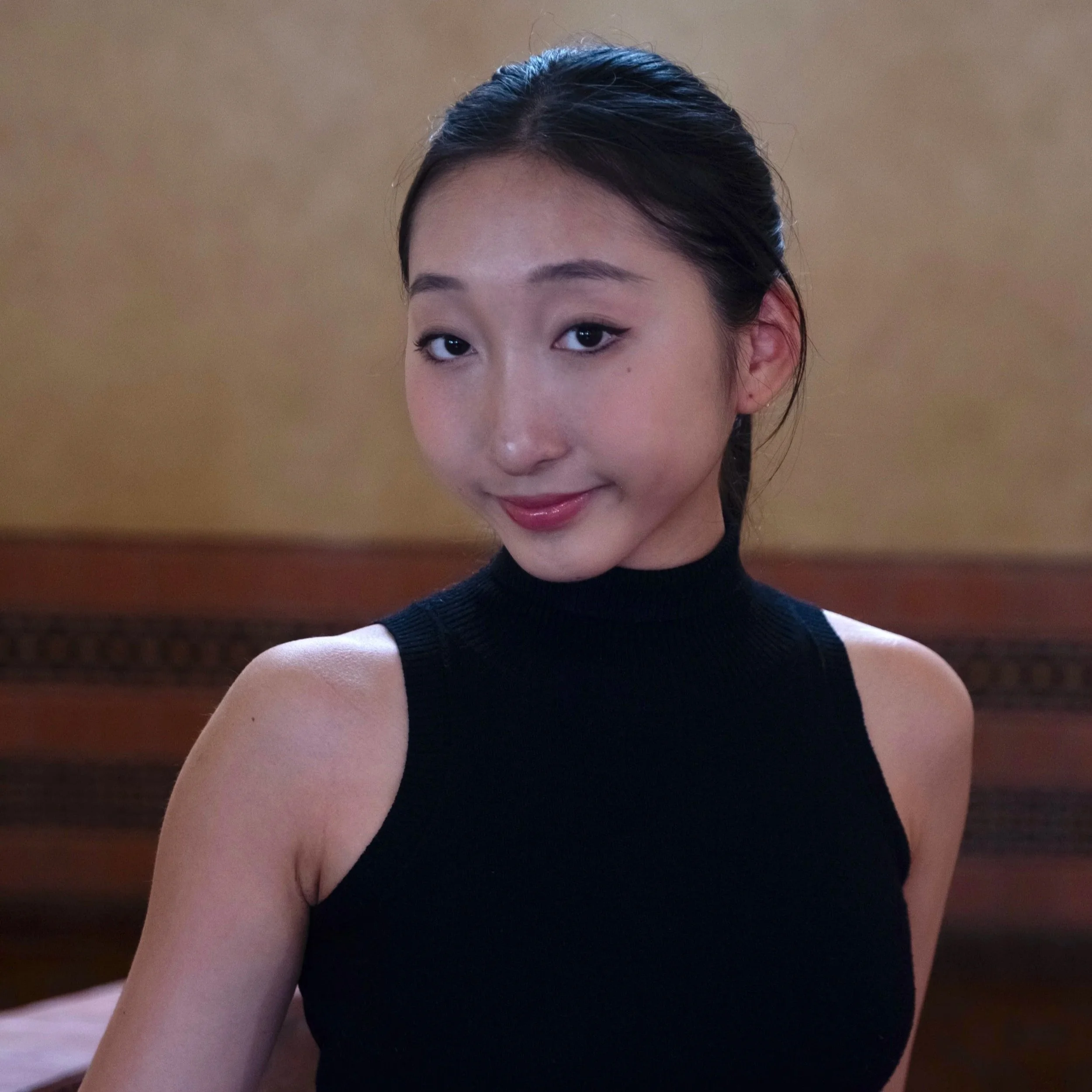 A young woman with dark hair pulled back, wearing a sleeveless black turtleneck top, smiling softly in an indoor setting with a warm-toned background.