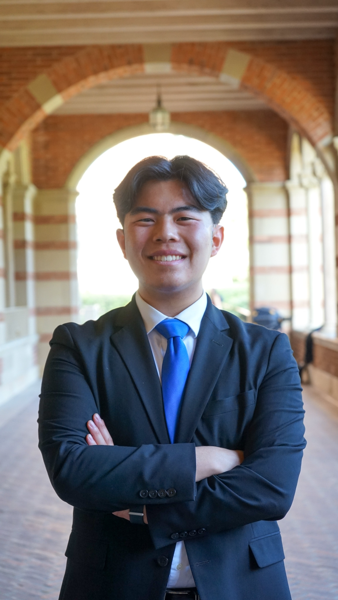 Young man in a black suit and blue tie smiling with arms crossed in front of an arched brick and stone building interior.