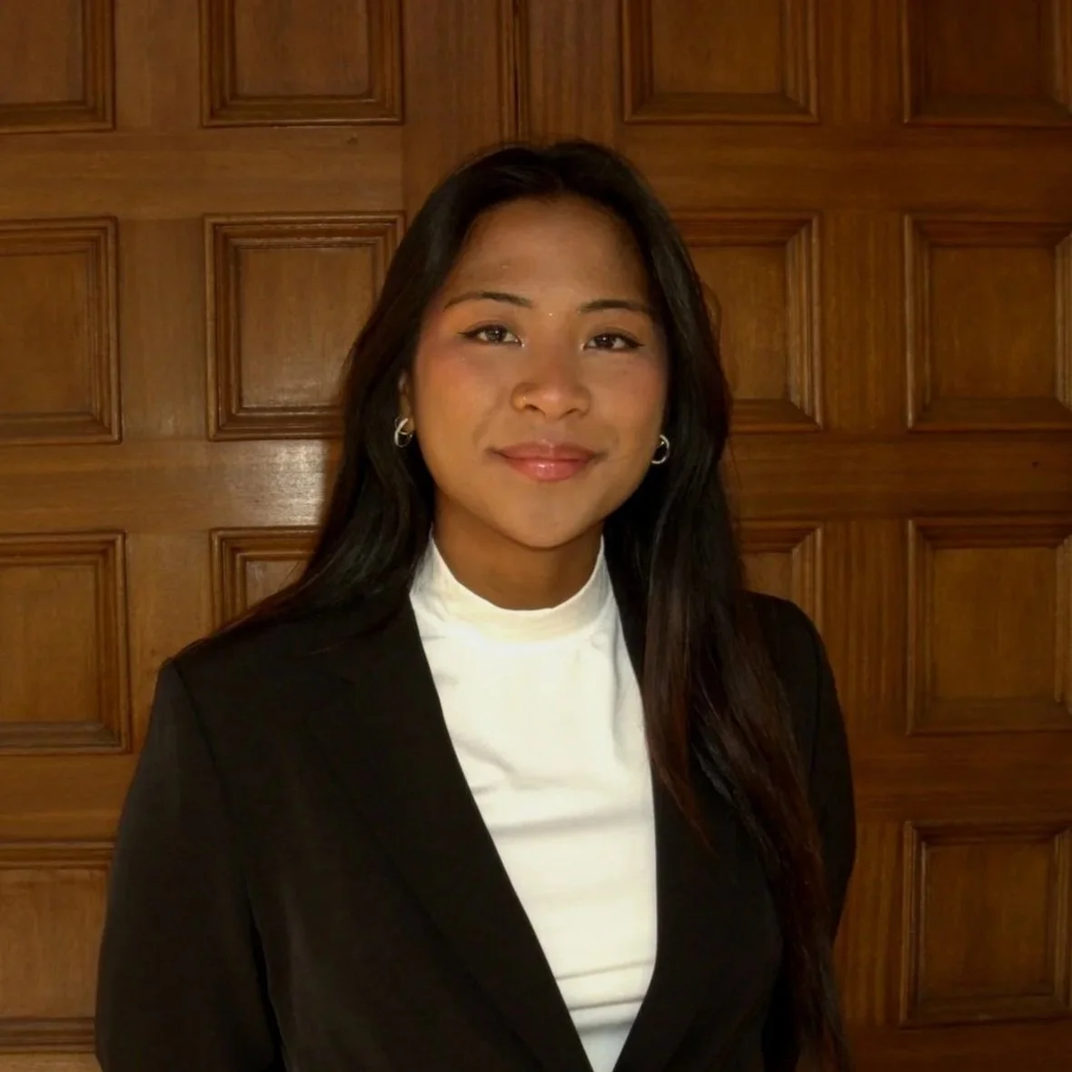A woman with long black hair, wearing a black blazer and white blouse, smiling in front of a wooden paneled background.