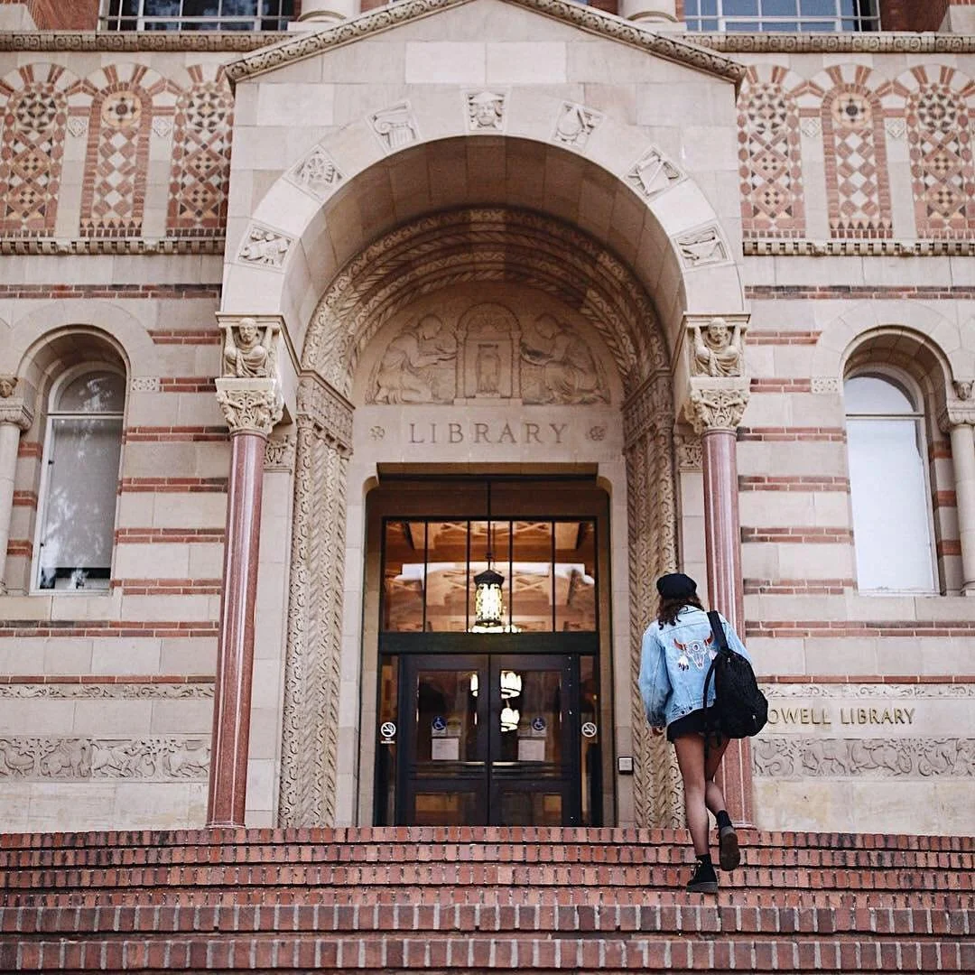 A person with a backpack, wearing a denim jacket and shorts, walking up the brick stairs into the UCLA Powell Library building with ornate architectural details and large windows.