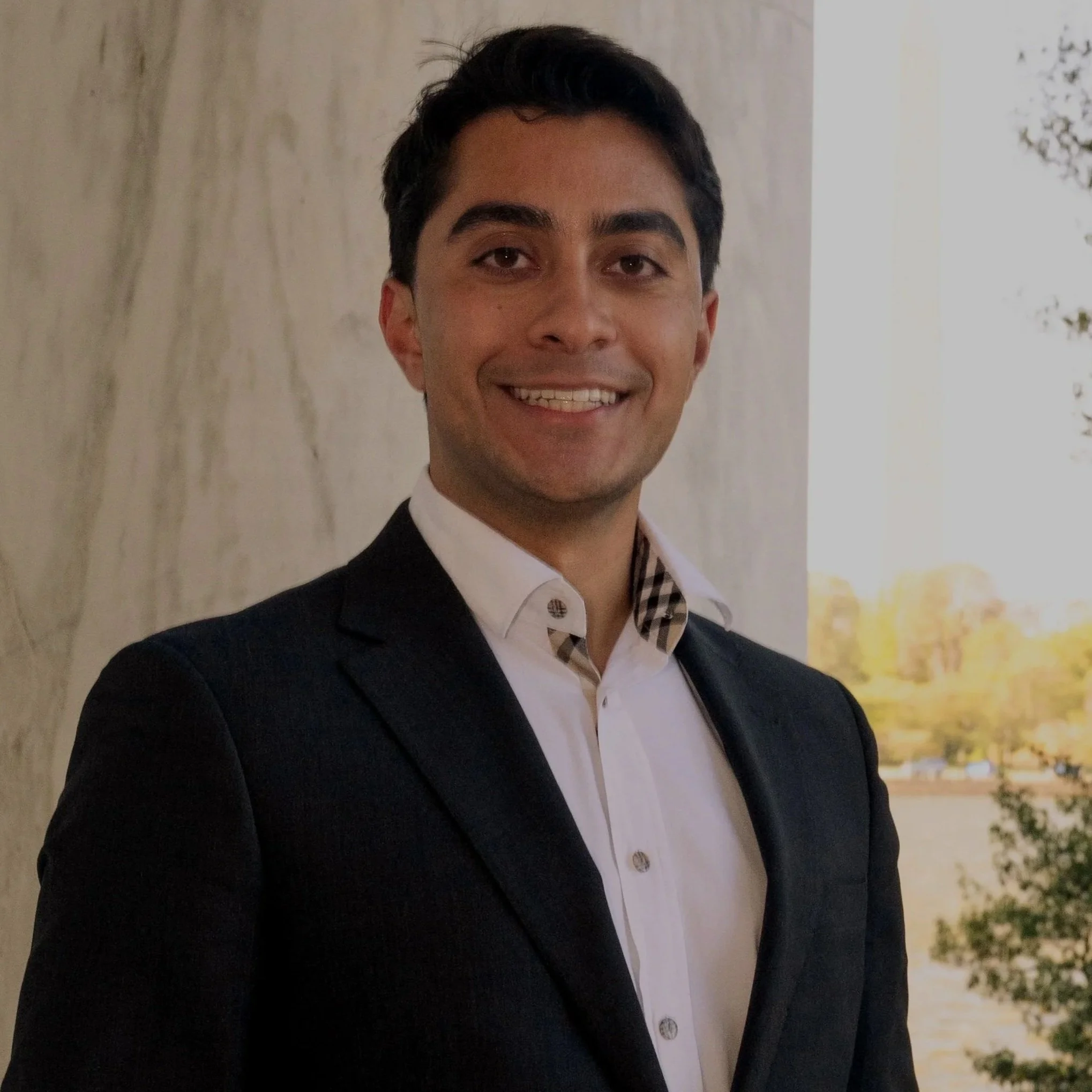 A man wearing a dark blazer and white shirt, smiling, standing outdoors with a blurred background of trees and water.
