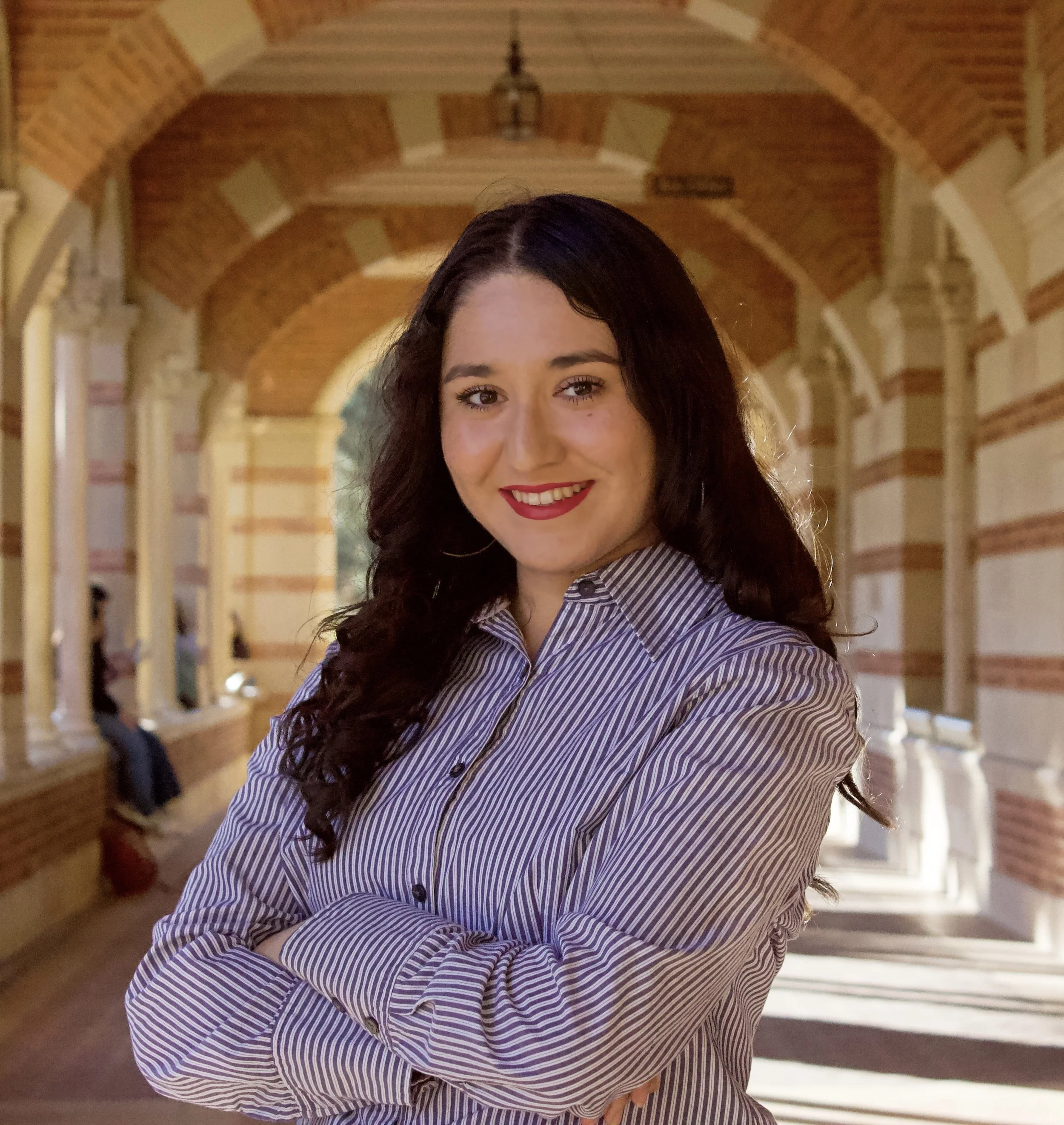A woman with long dark curly hair and red lipstick standing in a historic brick and stone corridor, smiling with arms crossed.
