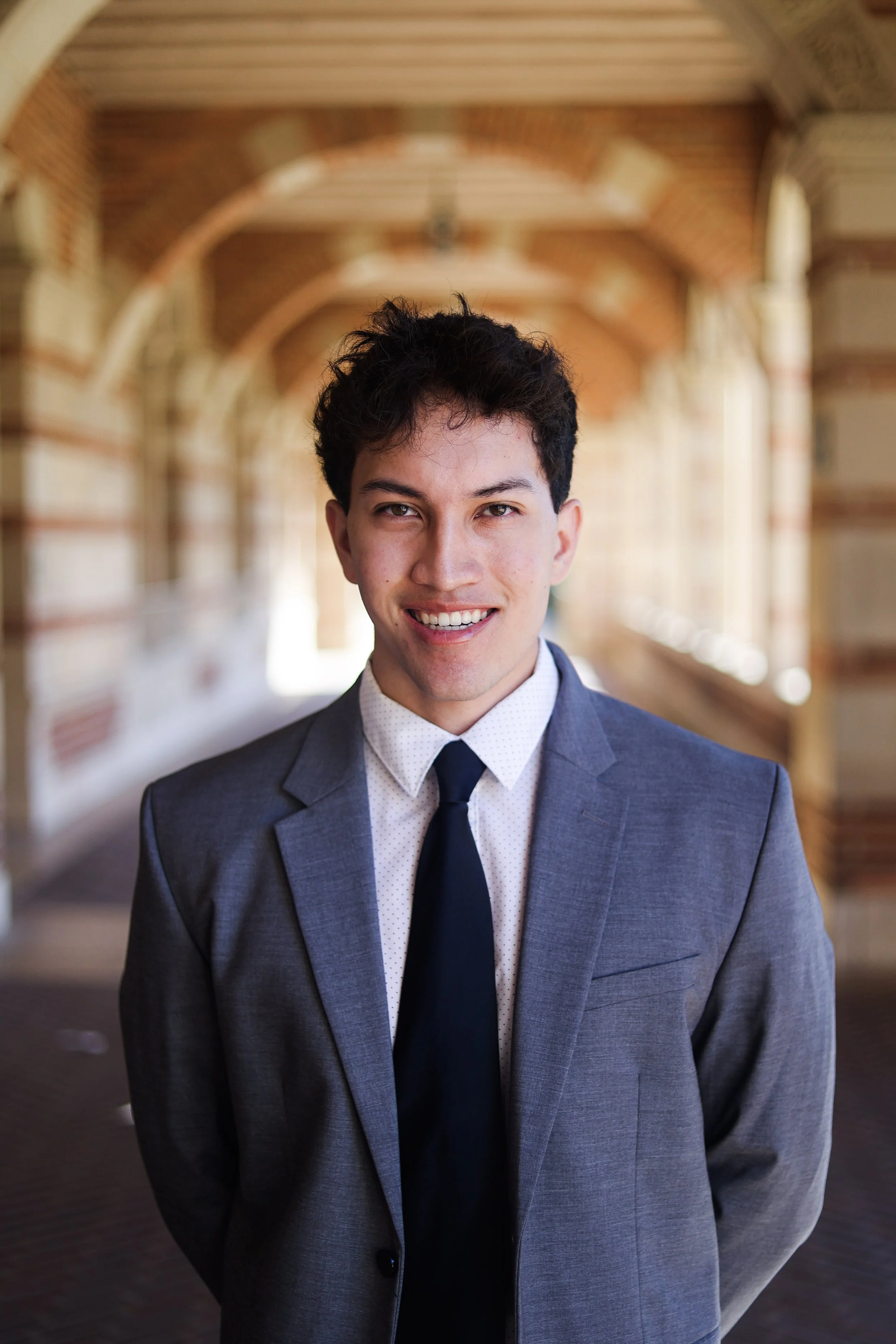 A young man in a gray suit, white shirt, and black tie standing outdoors in front of an arched brick and wood structure, smiling at the camera.