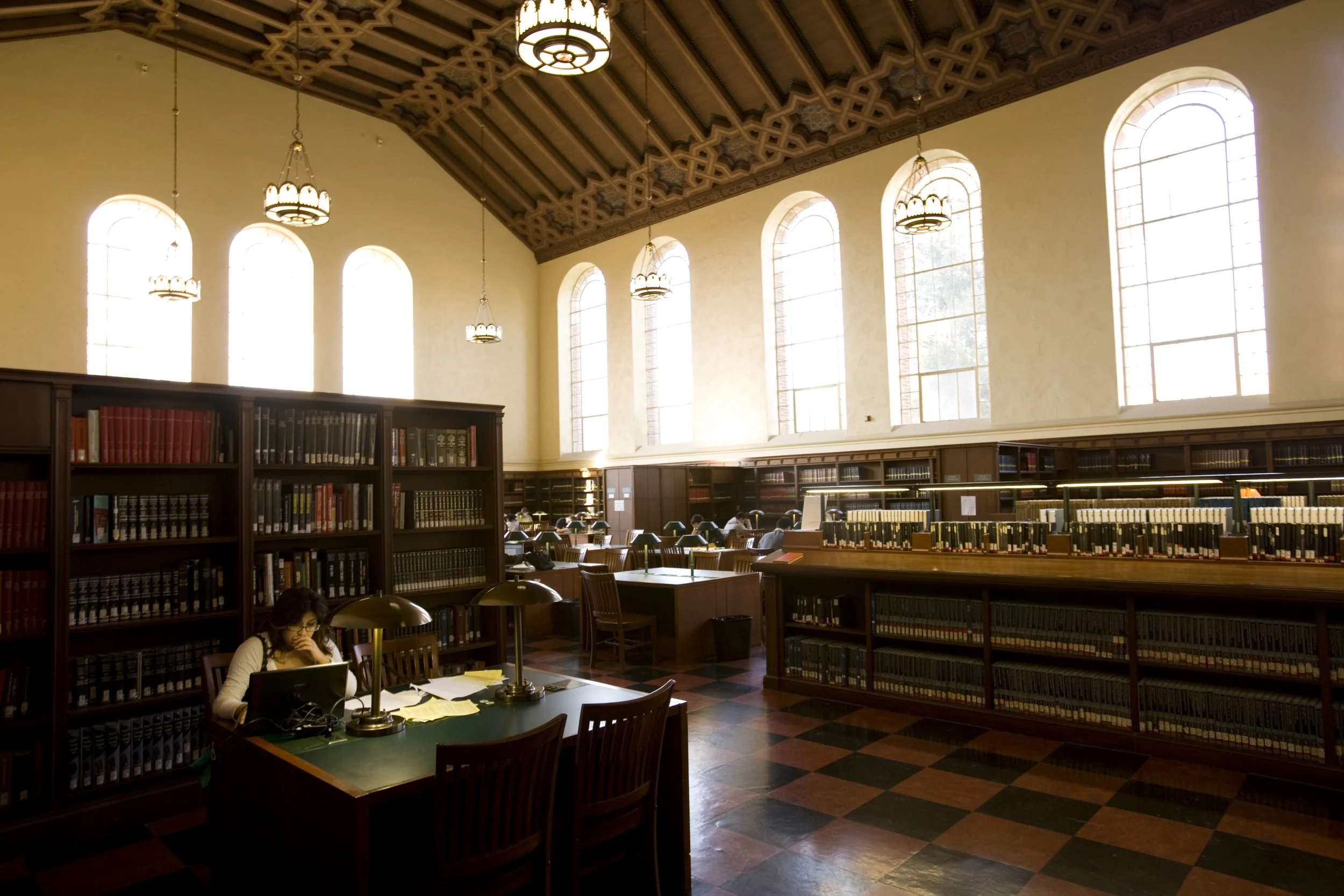 The Powell Library Reading Room, featuring tall arched windows, wooden bookshelves filled with books, and a reading area with tables and desk lamps. A person is sitting at one of the tables working on a laptop.