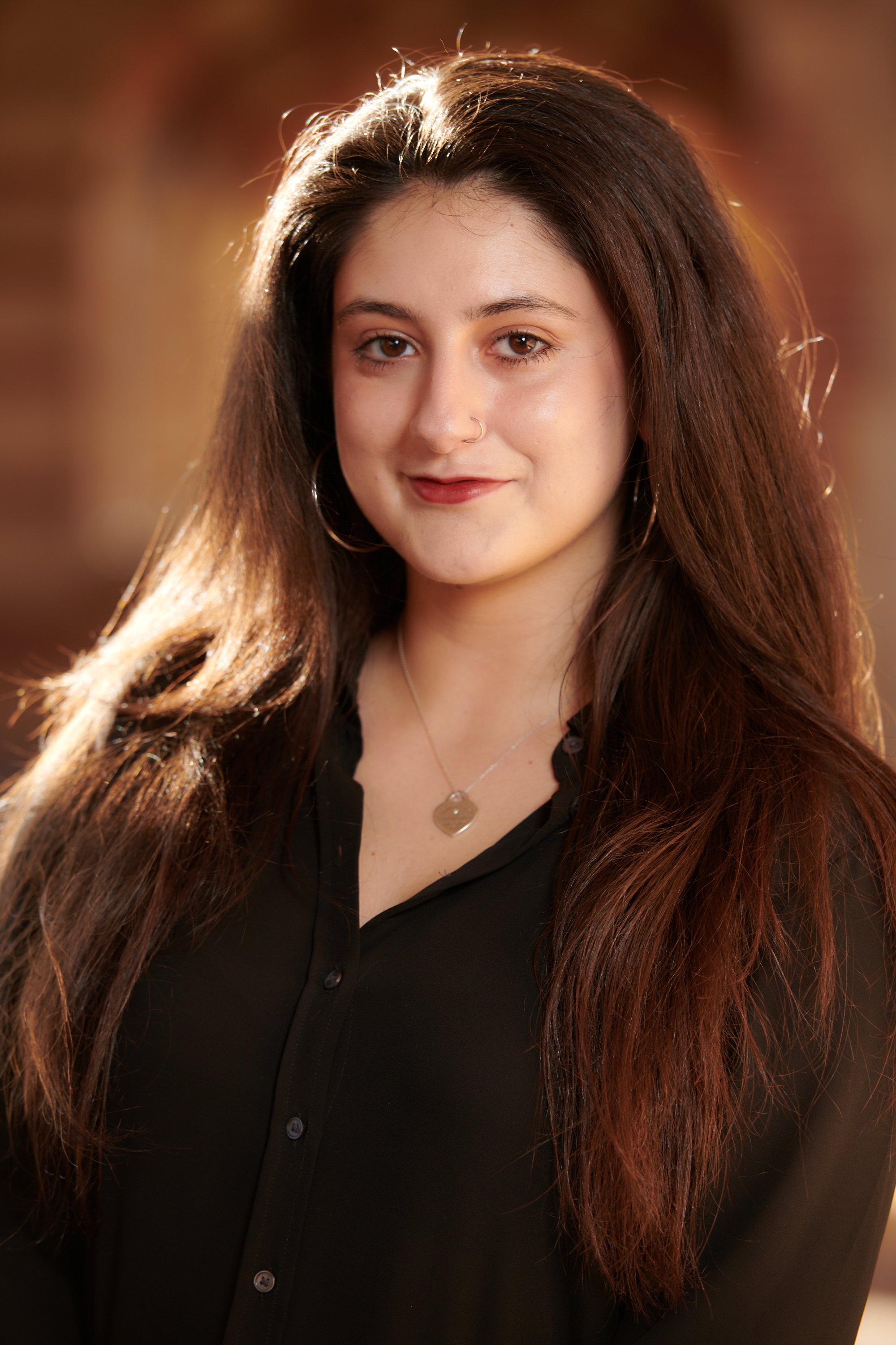 A young woman with long, brown hair, wearing a black shirt and a necklace with a round pendant, smiling softly at the camera with sunlight behind her.