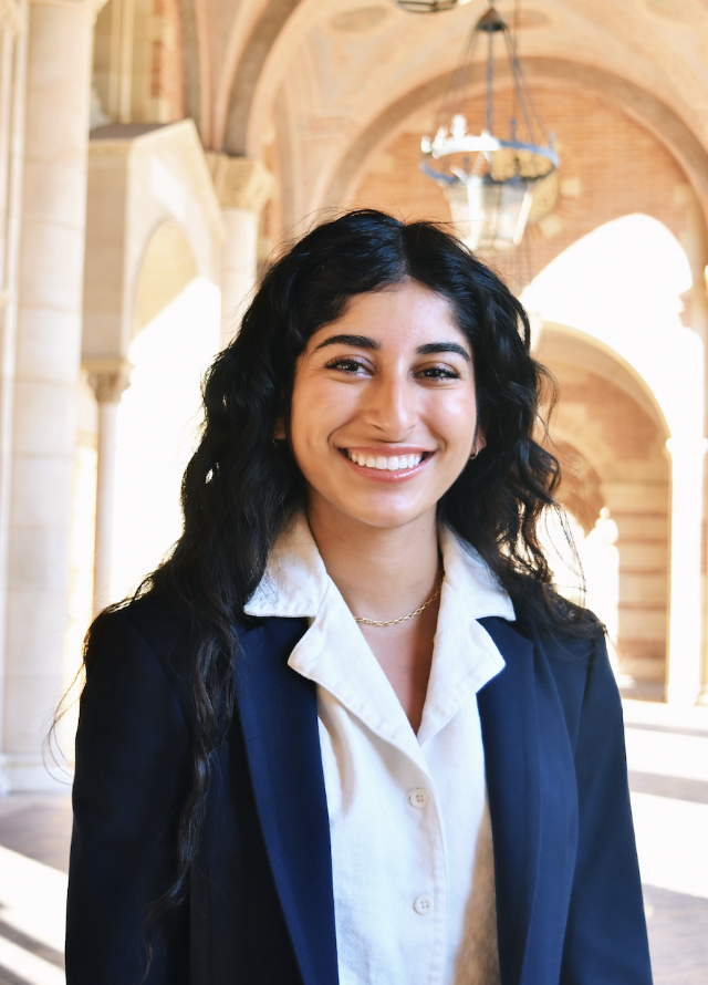 Young woman with long dark hair smiling, wearing a white blouse and dark blazer, standing in a building with arches and chandeliers.