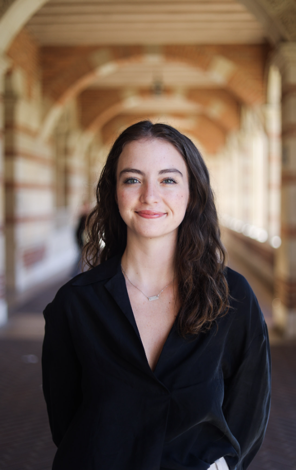 A young woman with brown, wavy hair and blue eyes, smiling, wearing a black blouse and a delicate necklace, standing in a covered outdoor walkway with brick and wood architecture.