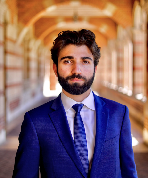 A man with dark hair and a beard, wearing a blue suit and tie, standing in an arched, brick corridor with sunlight streaming through windows.