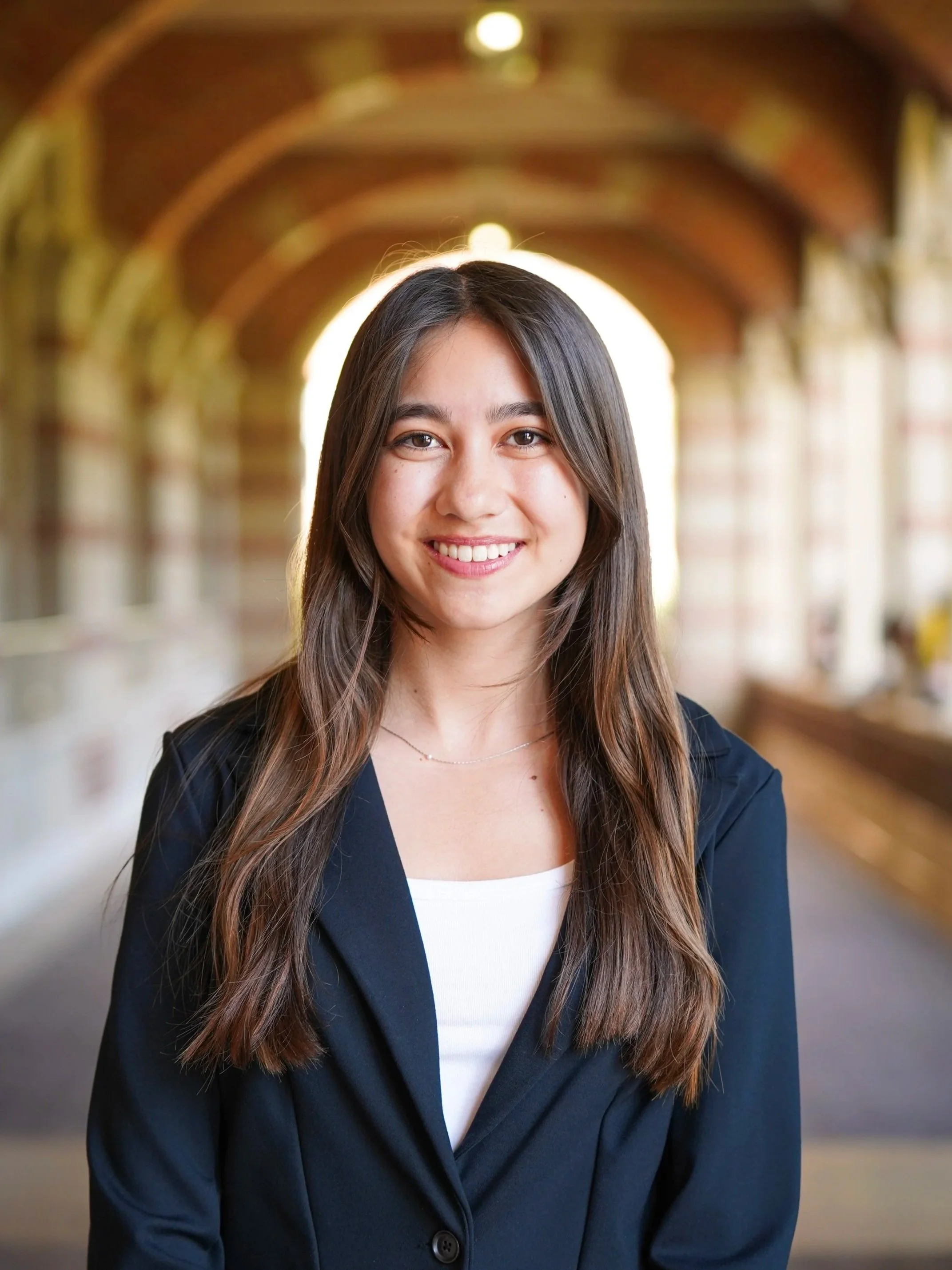 Smiling young woman with long brown hair wearing a navy blazer and white top, standing in a well-lit indoor corridor with brick archways and large windows.