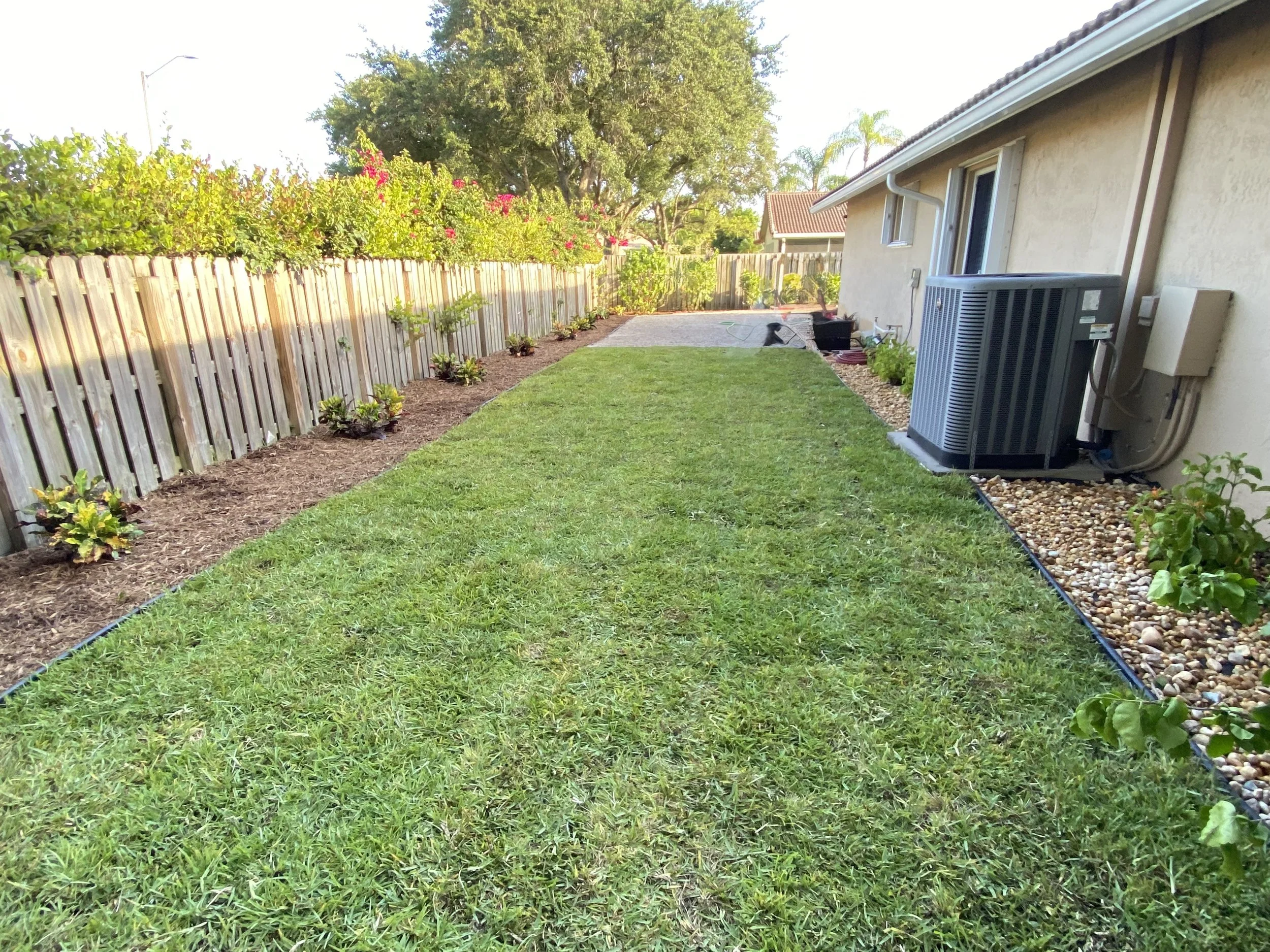 Backyard with green grass, flower bed with mulch along the house and fence, air conditioning unit, and potted plants near the house in bright sunlight.