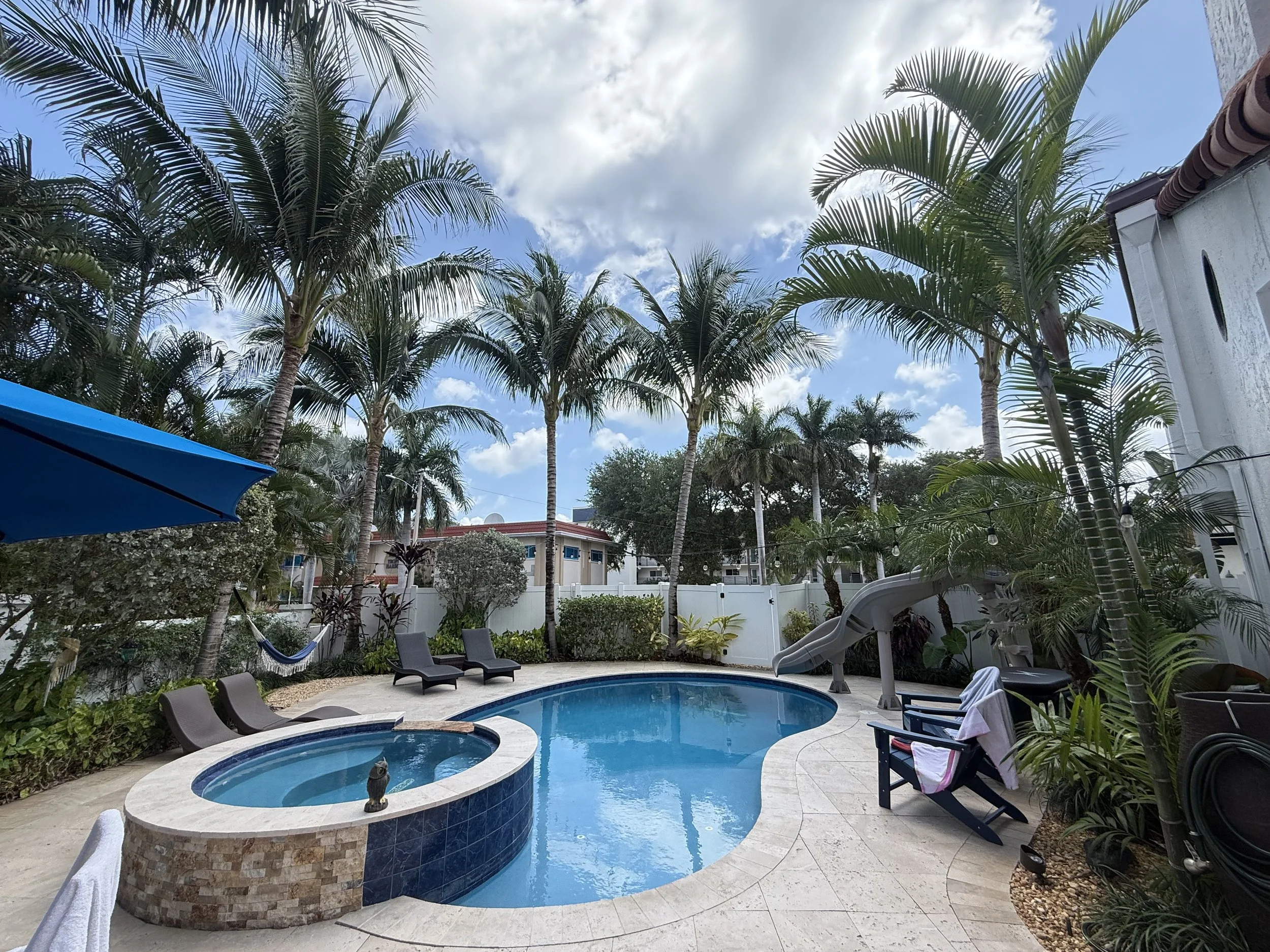 Backyard with swimming pool, hot tub, and palm trees. Features lounge chairs, a hammock, and a water slide. A blue umbrella and Adirondack chair with a towel are nearby.