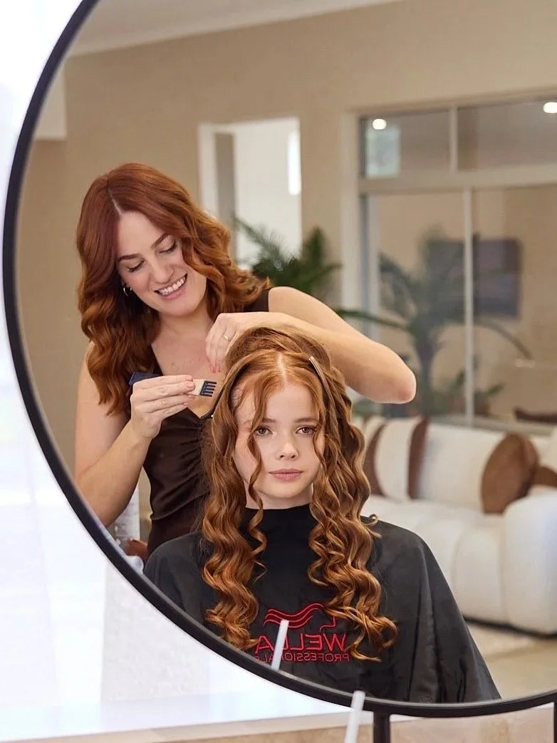 A woman with red hair and a white sleeveless top smiling at the camera, standing with her arms crossed.