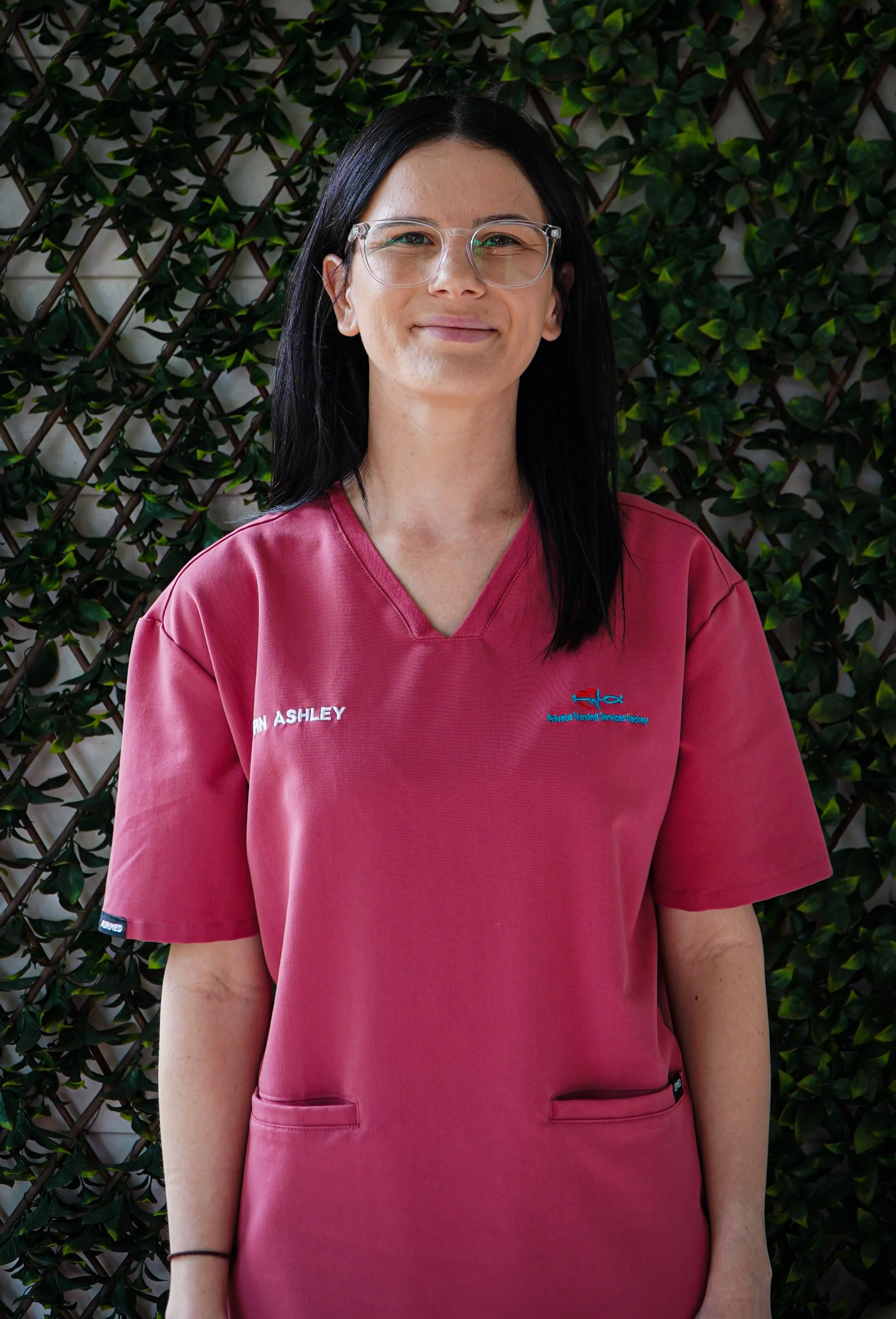 A woman with black hair, glasses, and a pink medical uniform, standing in front of a leafy green wall, smiling.