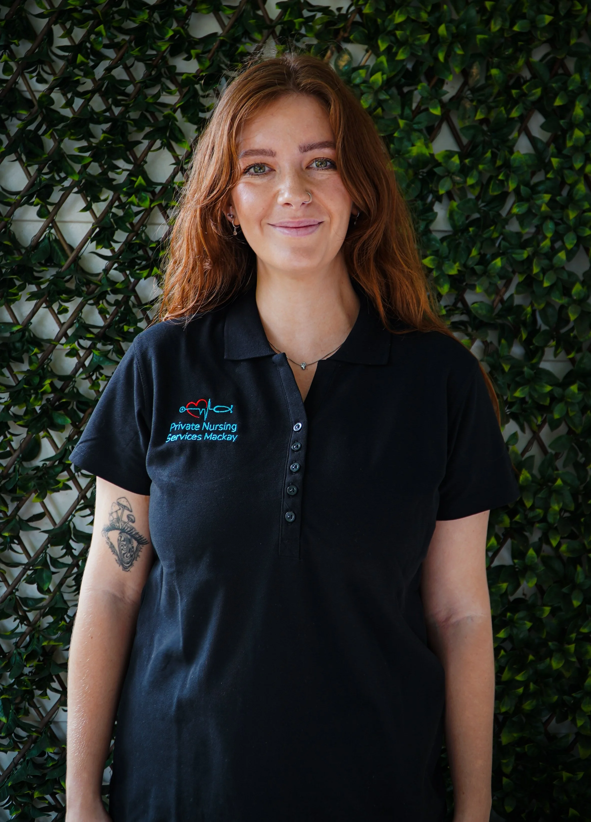 A woman with red hair wearing a black polo shirt with a logo and text that reads 'Private Nursing Services Mackay' standing in front of a green leafy background.