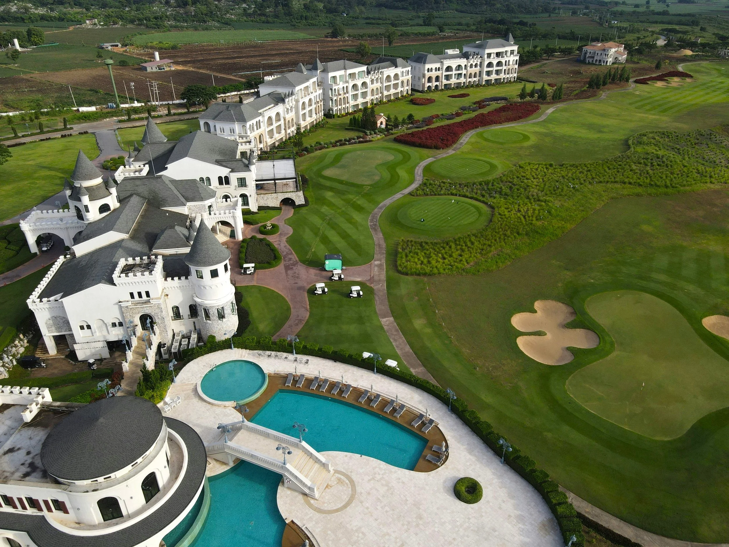 Aerial view of a large white castle-style building with multiple towers, a swimming pool, golf course, and landscaped green lawns surrounded by fields.