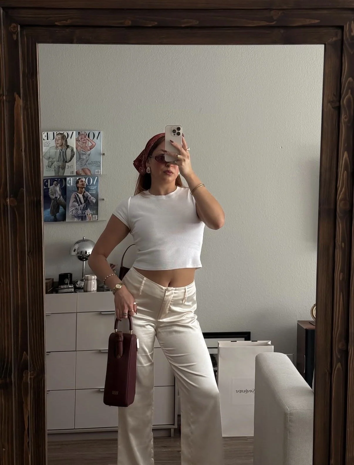 A woman in a purple dress with feathered shoulders sitting on a dark purple couch, holding a bottle of wine in her left hand, drinking wine from a glass in her right hand, in a room with framed artwork on the wall and a white lamp on a wooden side table.