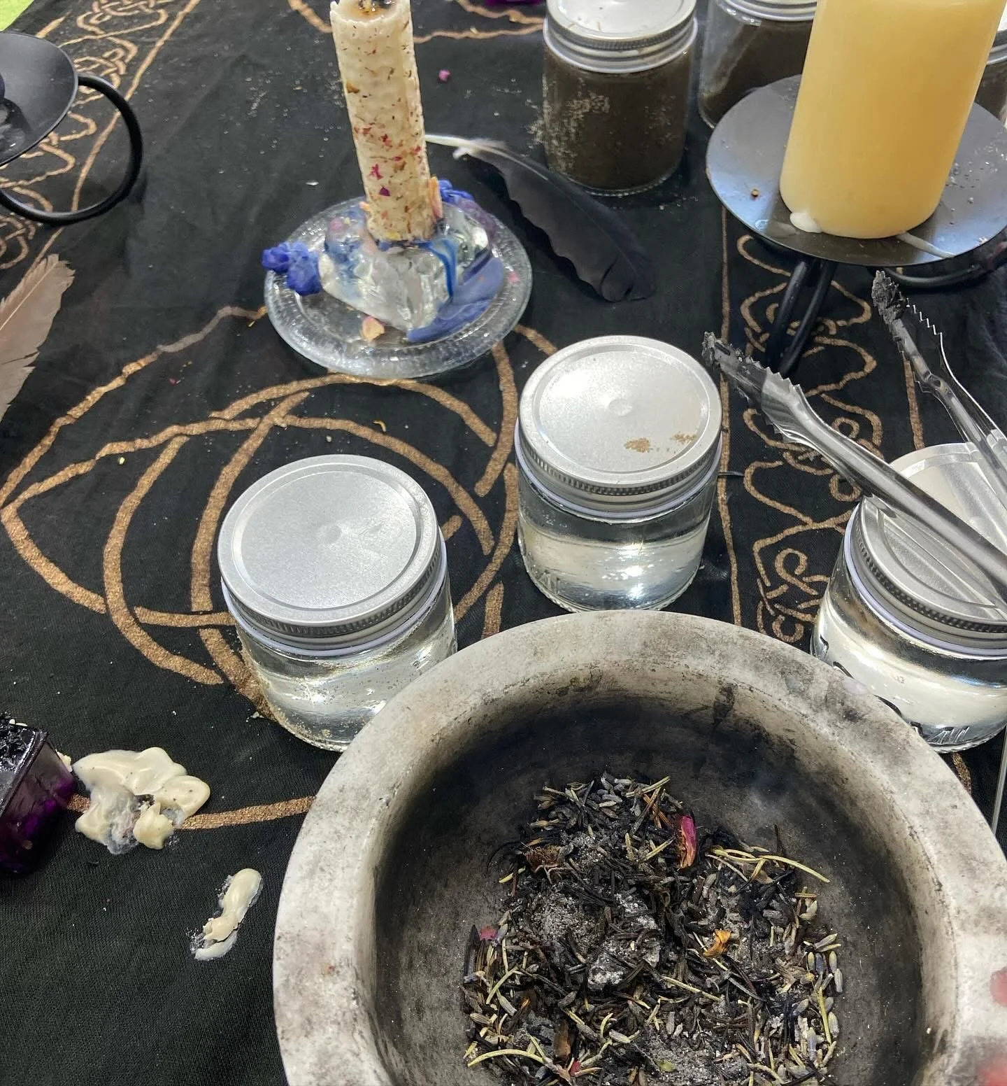 A table with ritual items, including a large candle, three jars of water, a mortar with burnt herbs, and various candles and objects, set on a black cloth with symbolic gold patterns.