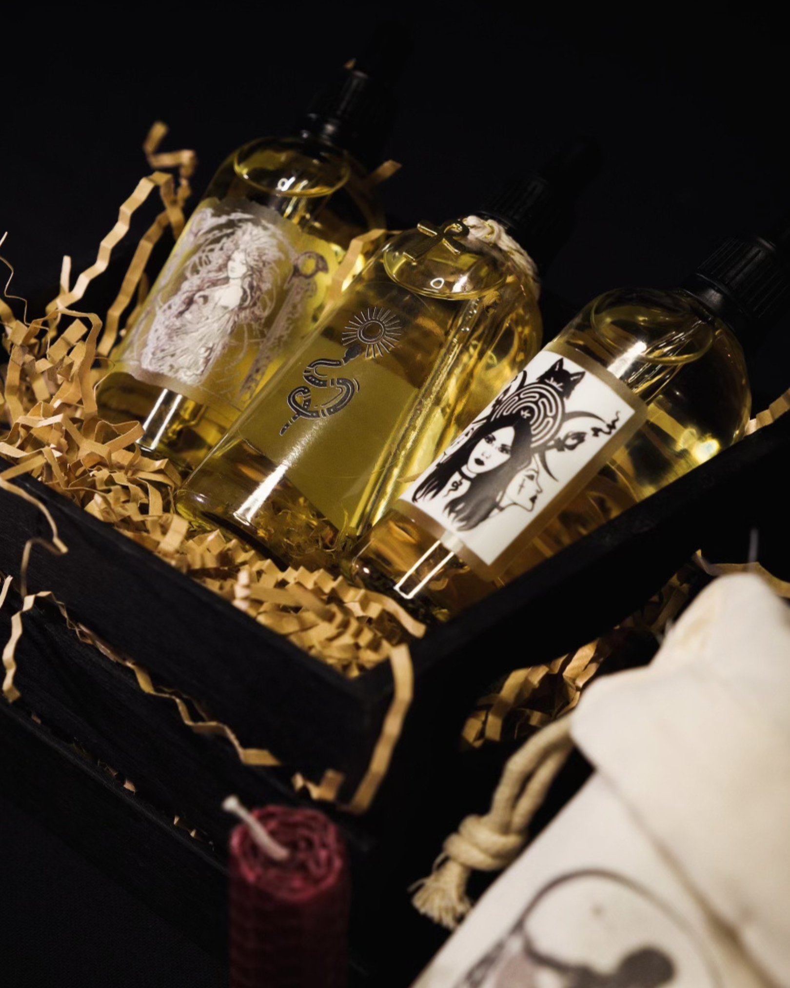 Three perfume bottles with black spray tops in a black box with shredded paper, dark background, and a partially visible white bag and red candle in the foreground.