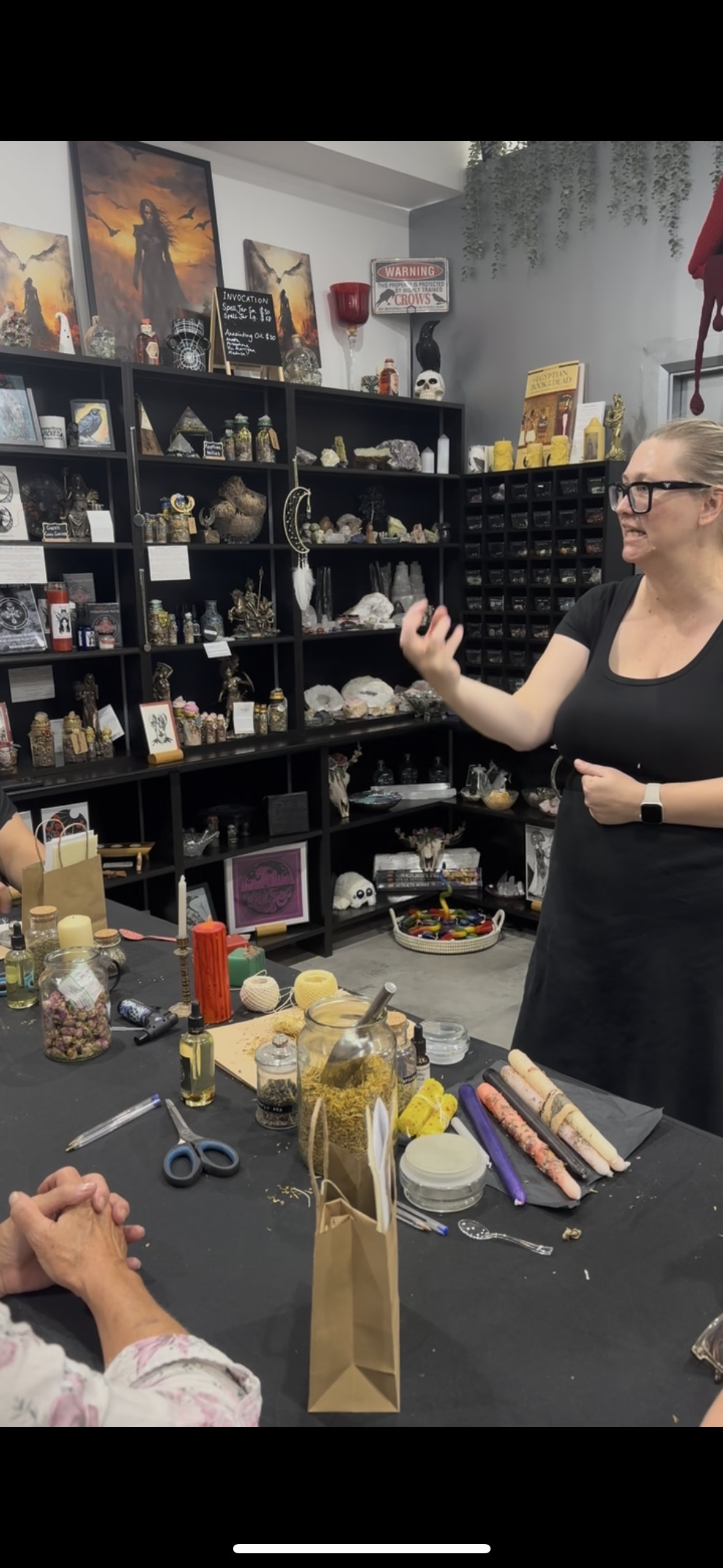 A woman with glasses and a black shirt stands at a workspace in a craft or metaphysical store, gesturing with her hand while people sit at the table. The table is cluttered with crafting supplies such as jars, candles, scissors, and stones. Shelves behind her display various mystical items, including artwork, skulls, crystals, and decorative objects.