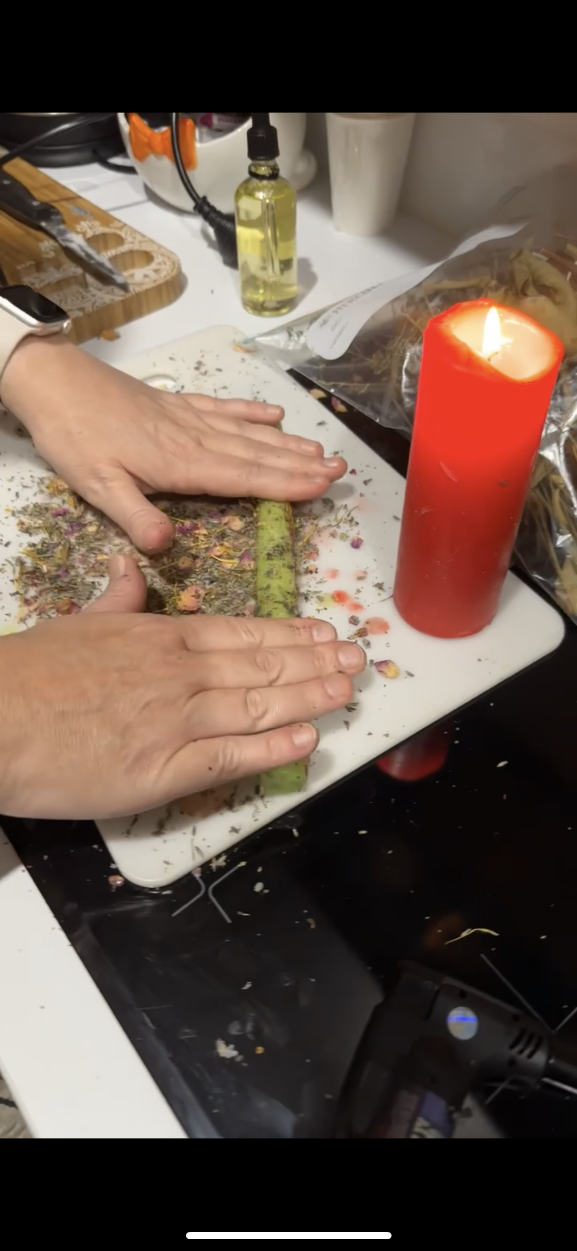 Person chopping dried flower petals on a white cutting board, with a lit red candle nearby, in a cluttered kitchen or workspace.