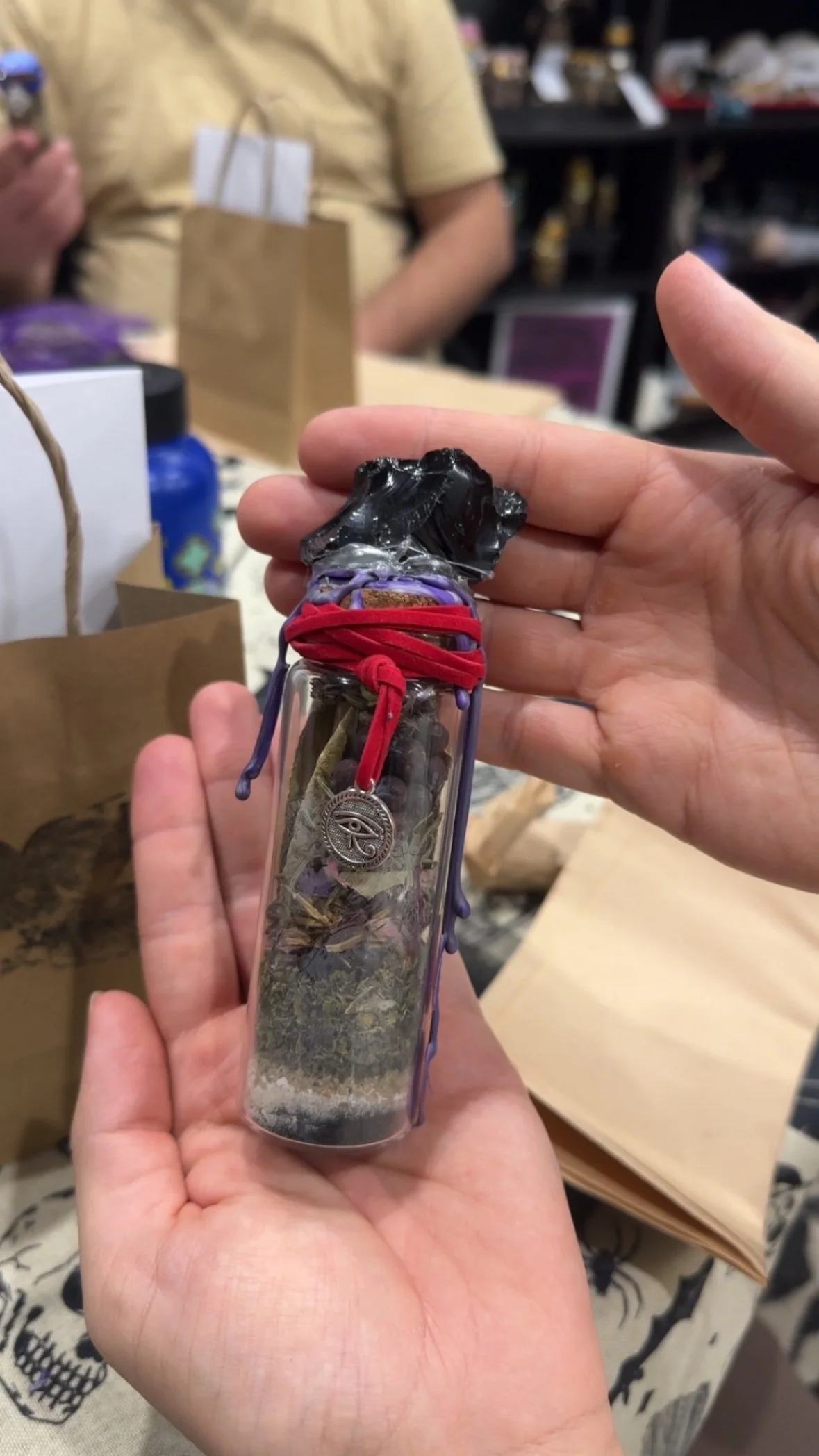 A hand holding a glass jar filled with herbs, with a black plastic bag sealed on top, adorned with red and purple strings, and a small pendant hanging from the strings.
